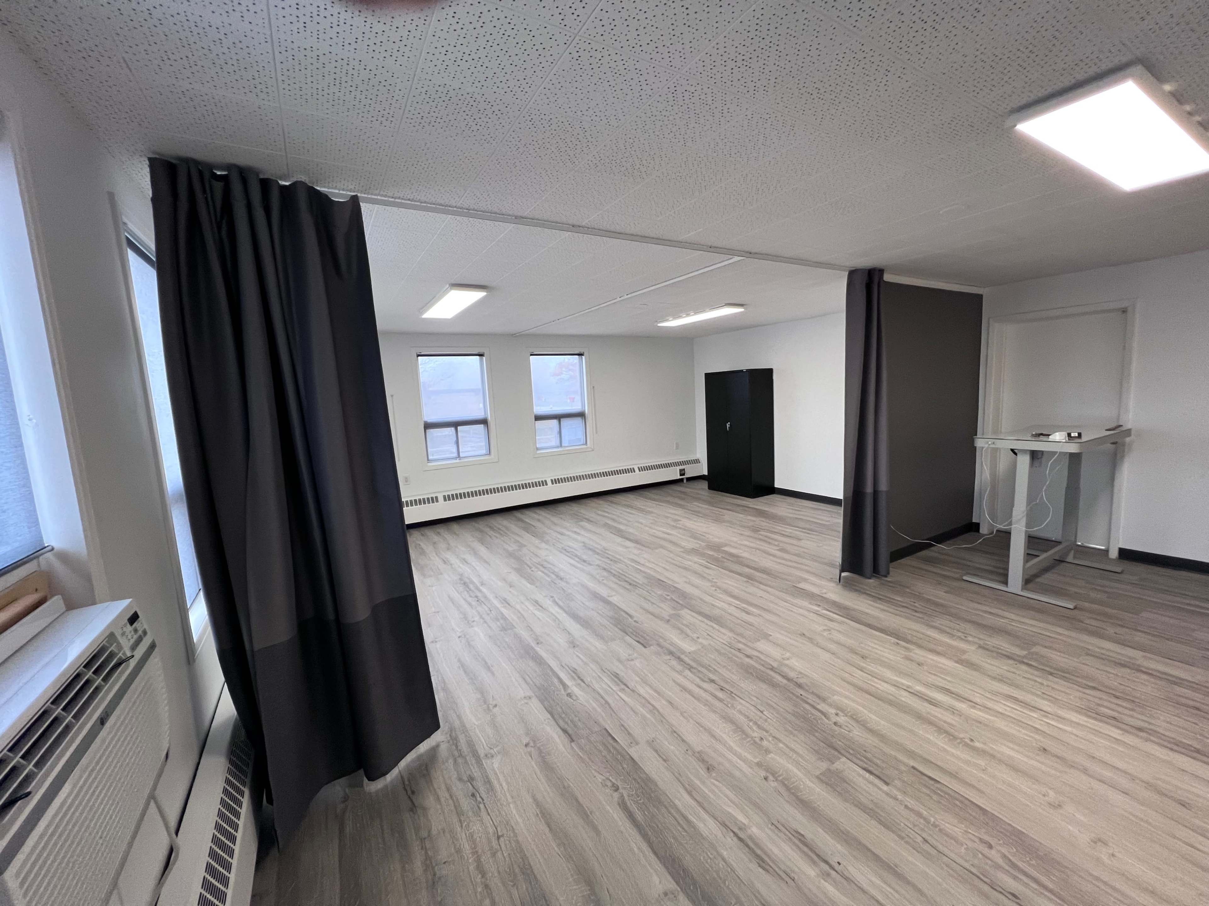 A spacious, empty room with gray curtains, a standing desk, and large windows that allow natural light.