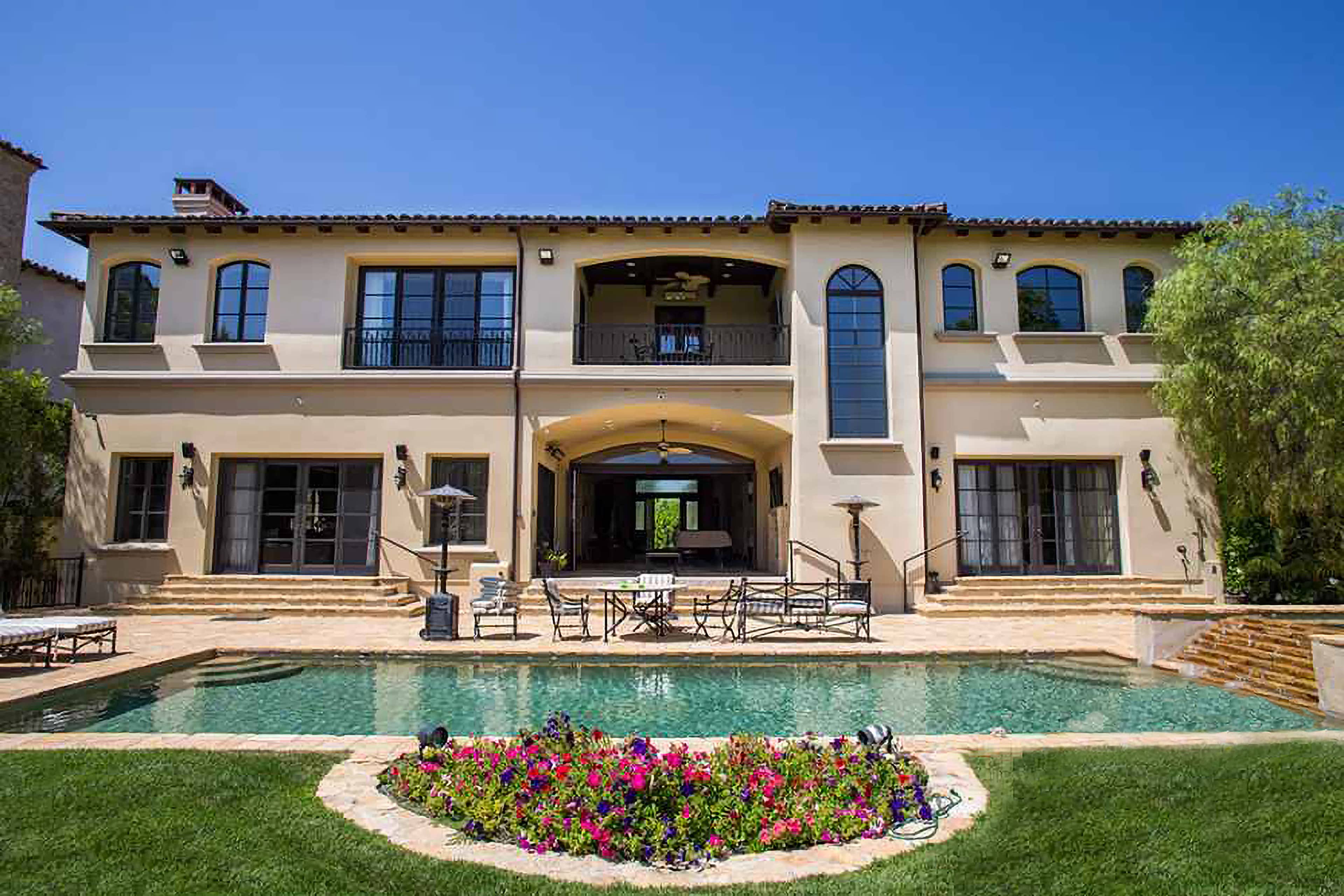 A two-story Mediterranean-style house with a spacious backyard, featuring a swimming pool surrounded by vibrant flowers and outdoor seating.
