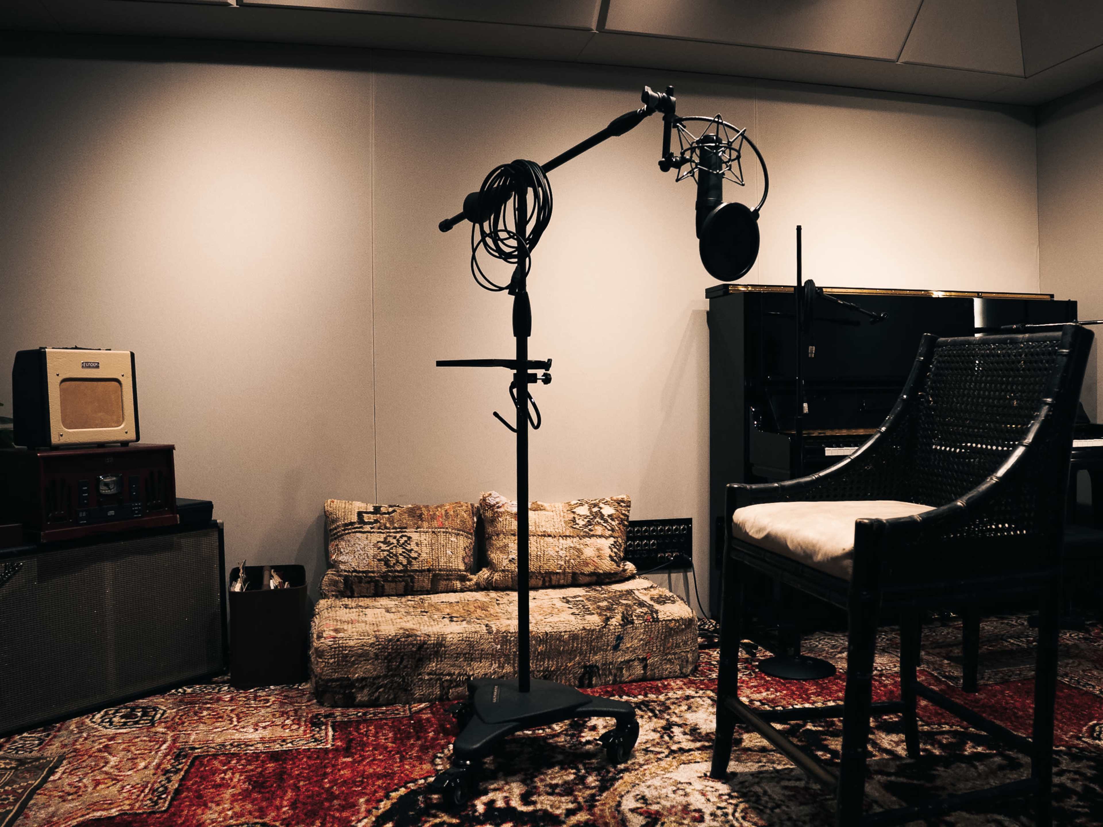 The image shows a sound studio featuring a microphone on a stand, a chair, a vintage radio, and a piano against a wall with acoustic panels.
