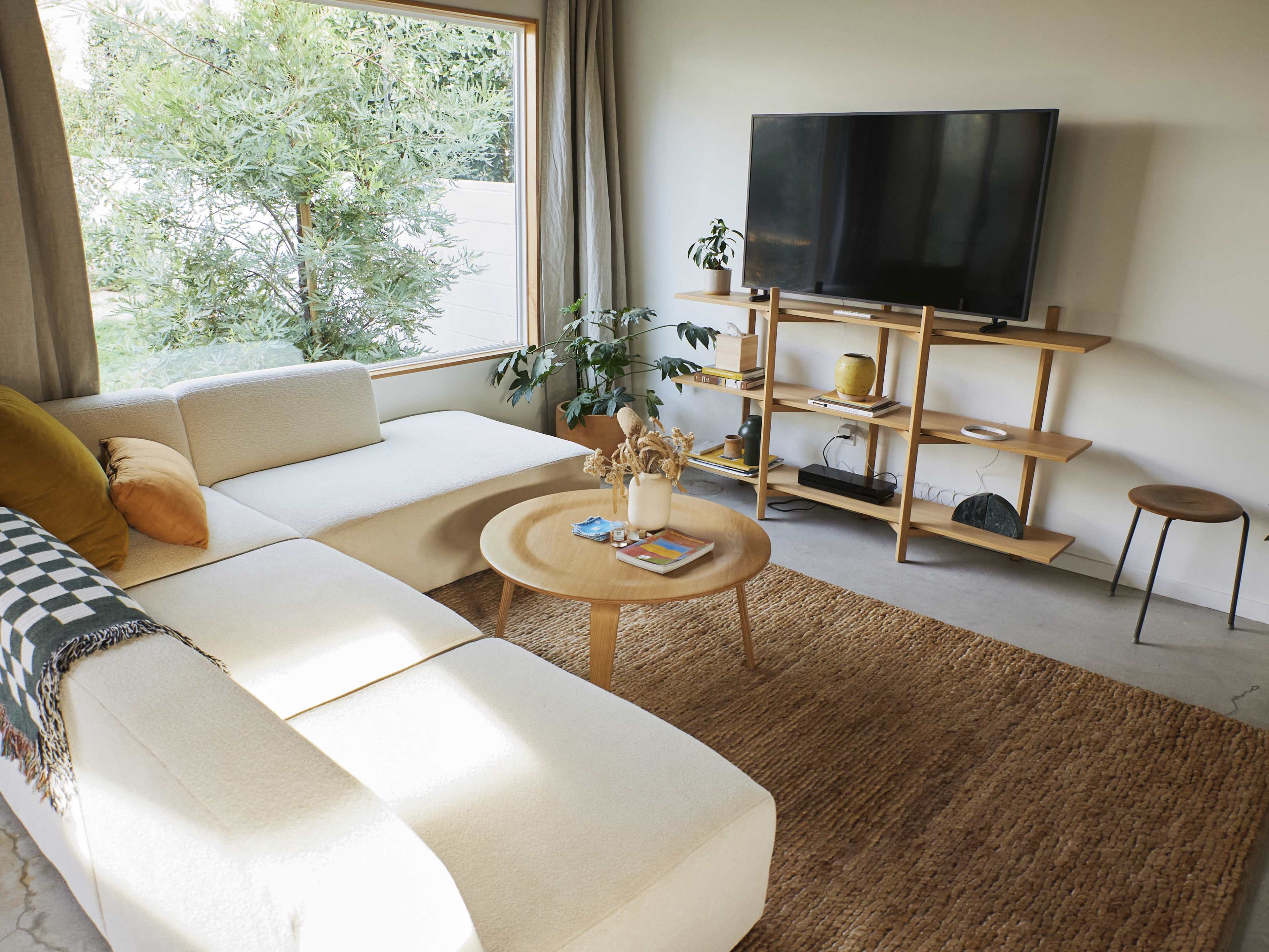 A modern living room features a light-colored sectional sofa, a round wooden coffee table, a television on a shelving unit, and large windows providing natural light.