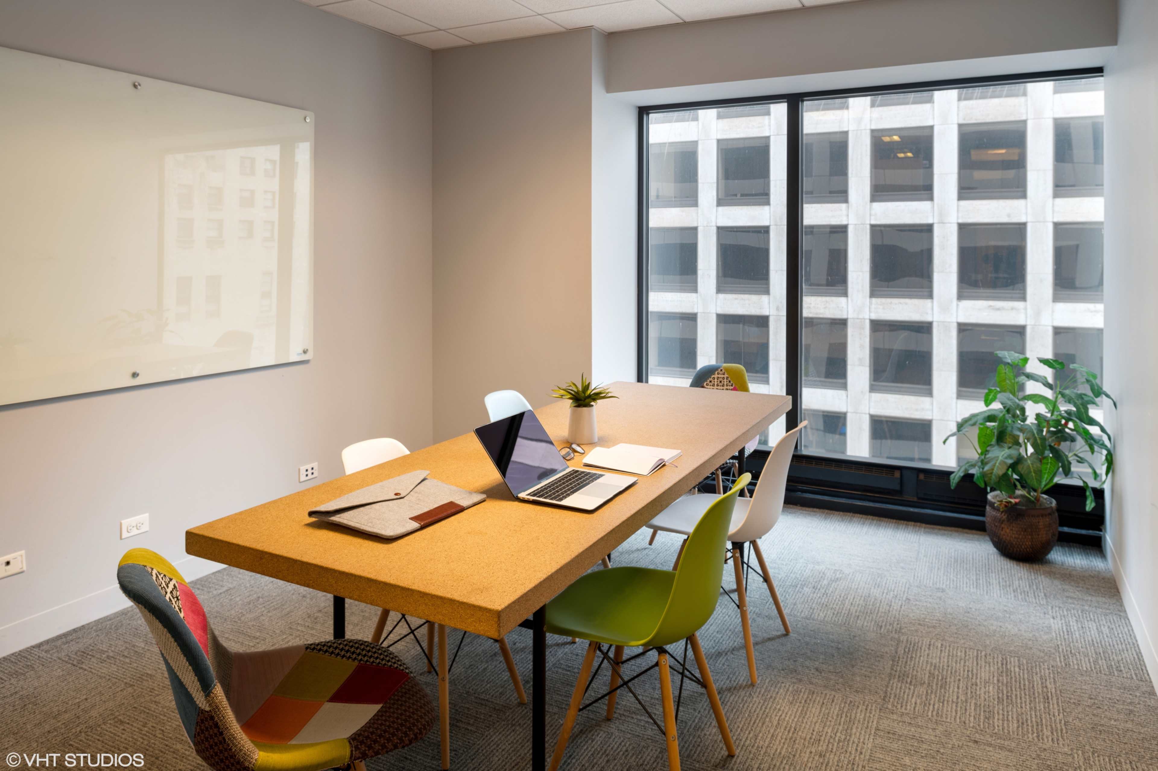 A modern conference room features a large cork table surrounded by colorful chairs, with a laptop and notepad on the table and a view of a building through the window.