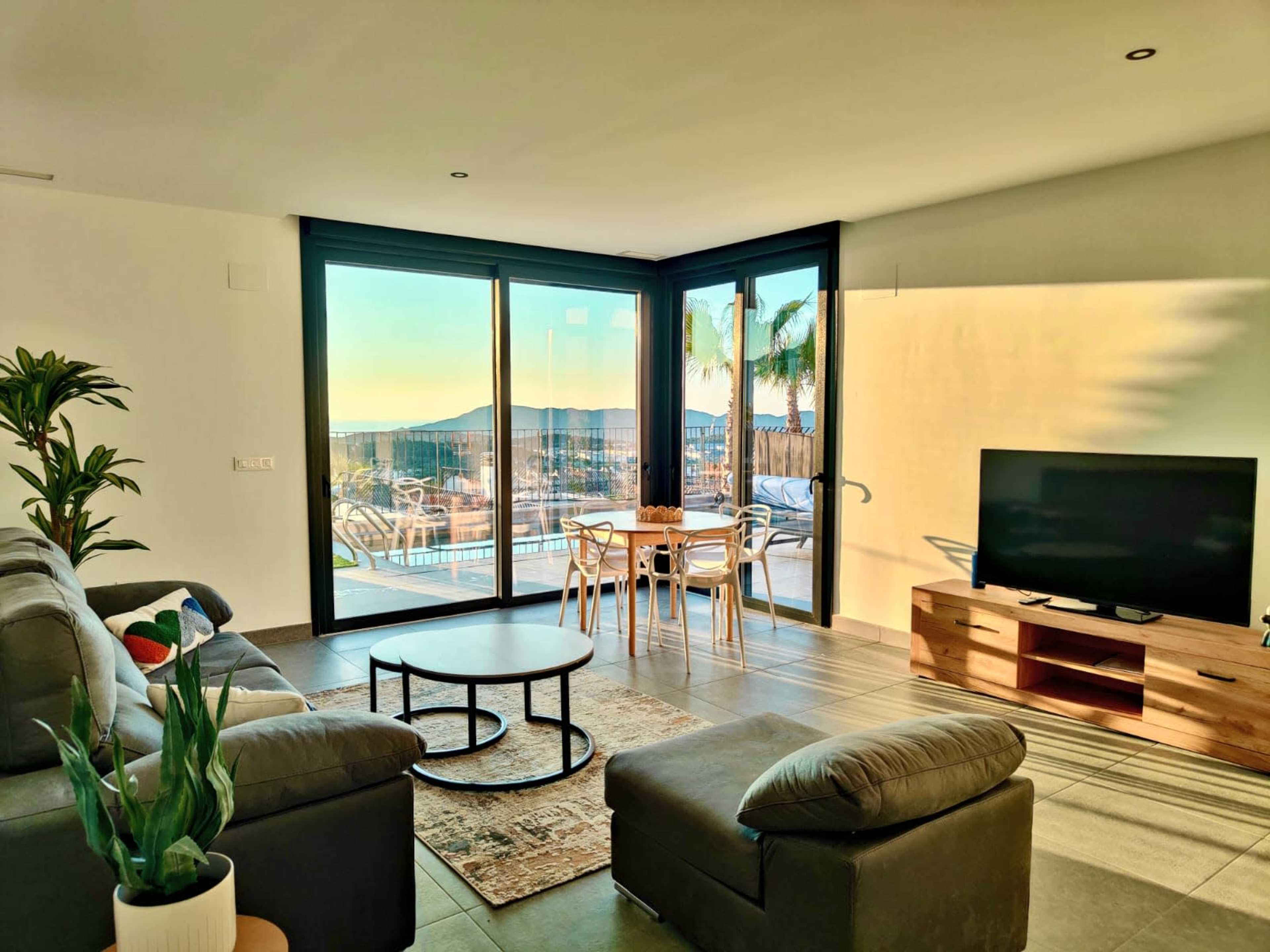 A modern living room features a sofa, television, and dining area with large windows showcasing a view of an outdoor pool and distant mountains.