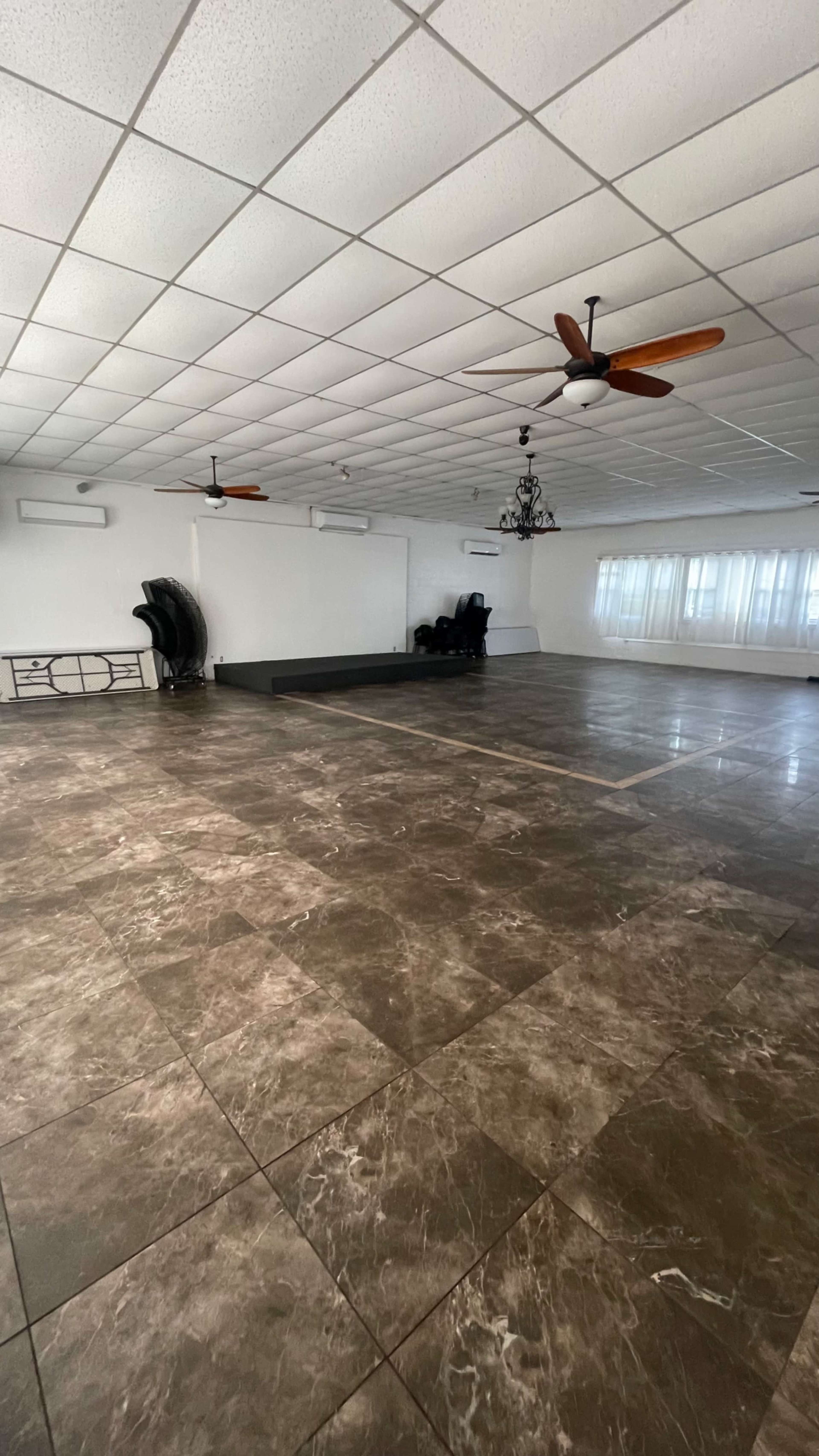 The image shows a spacious room with a polished tile floor, ceiling fans, and several chairs arranged along the wall.