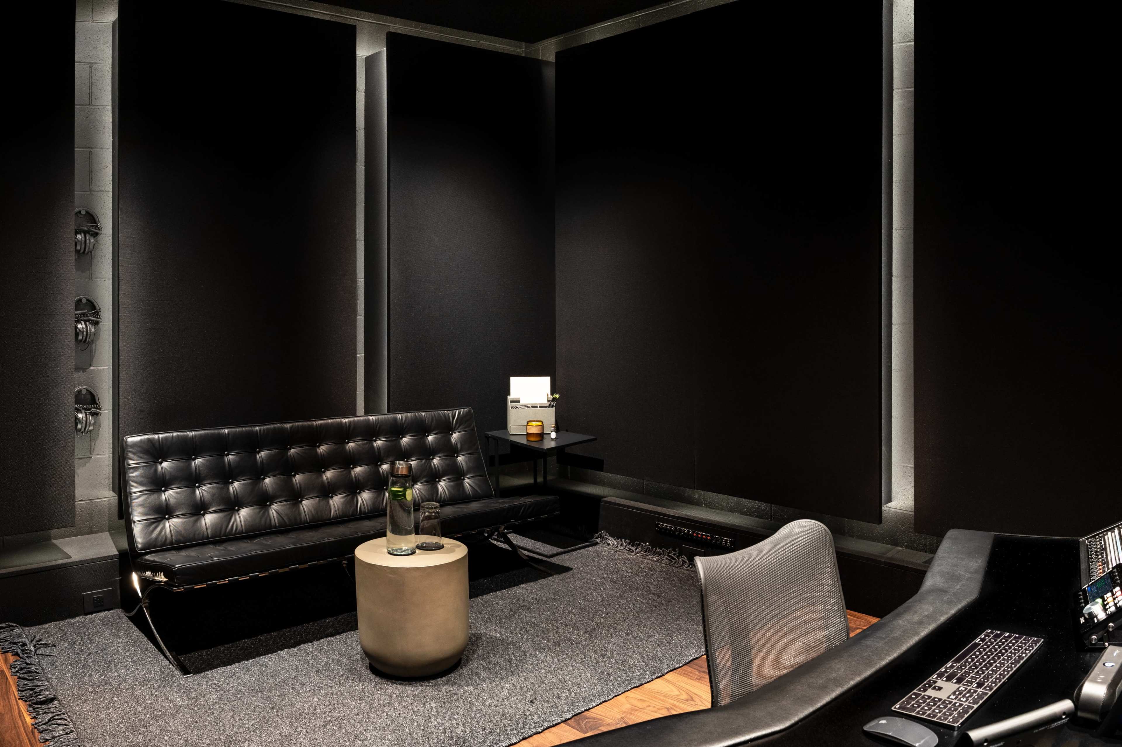 The image shows a modern, dimly lit room with a black tufted sofa, a round stone coffee table, and a sleek desk with audio equipment.