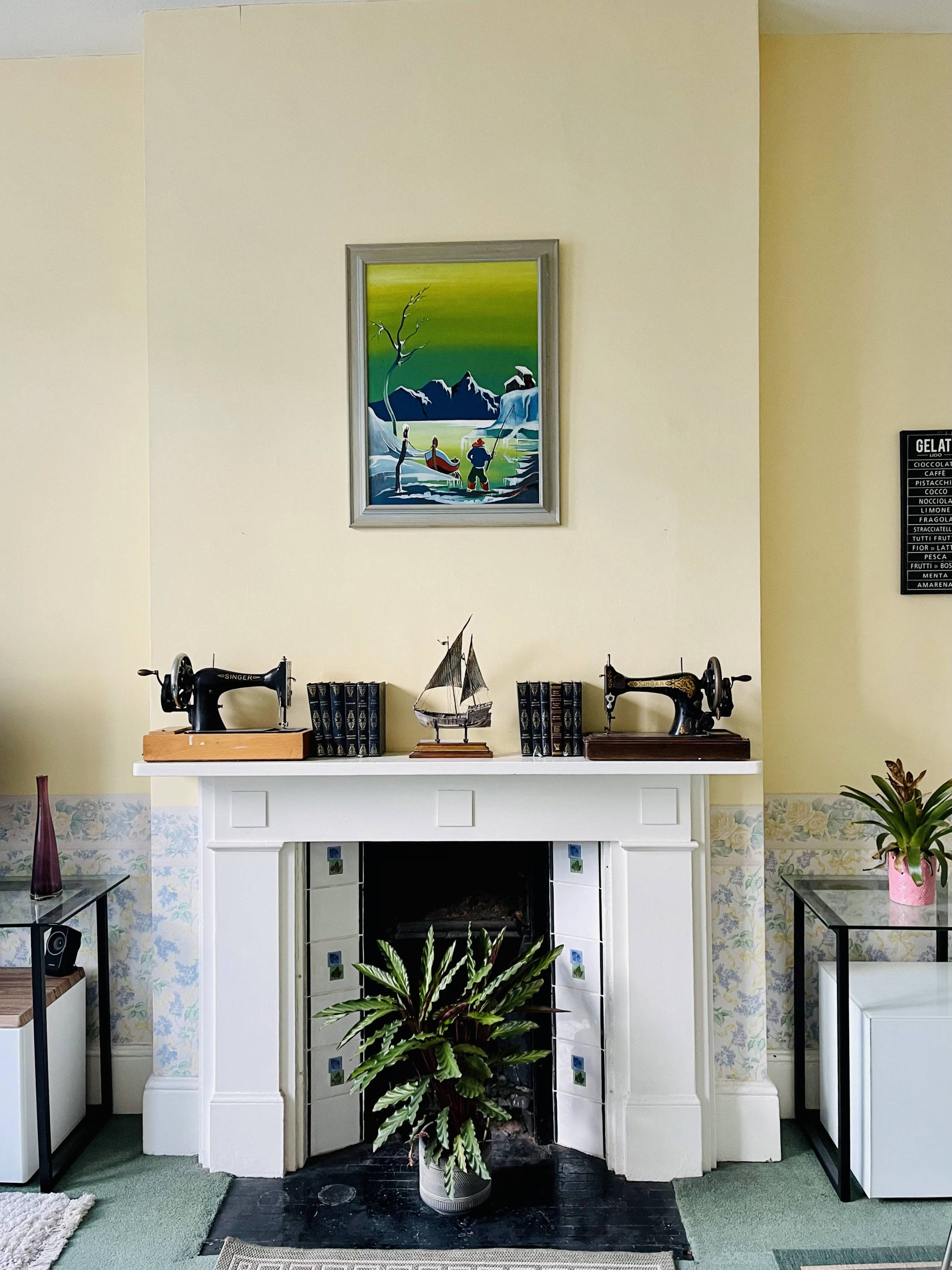 A mantelpiece with two vintage sewing machines flanking a decorative sailboat model and a painting of a landscape hanging above it, set against a light yellow wall.