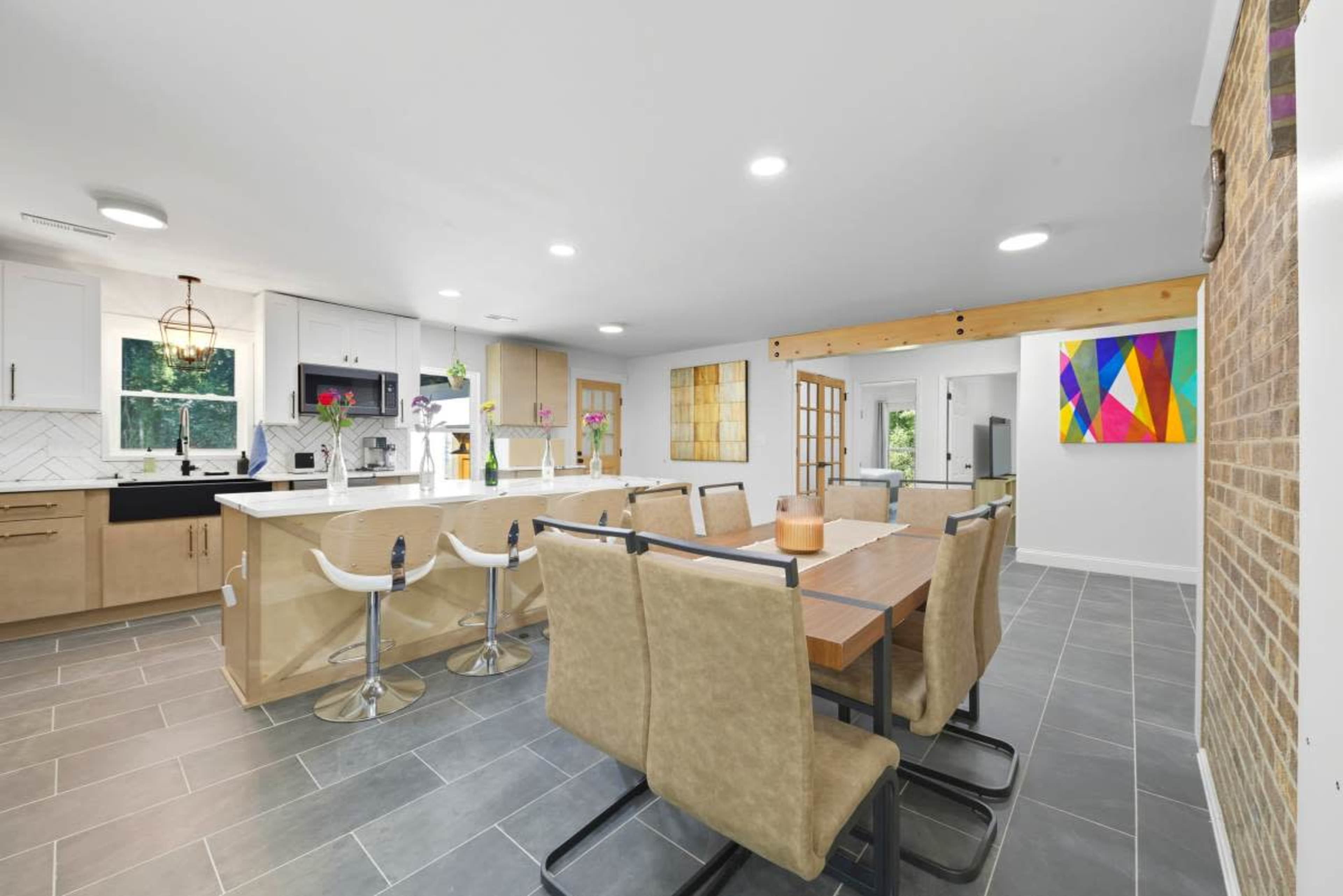 The image shows a modern kitchen and dining area with a long wooden table, bar stools at an island, and colorful artwork on the walls.