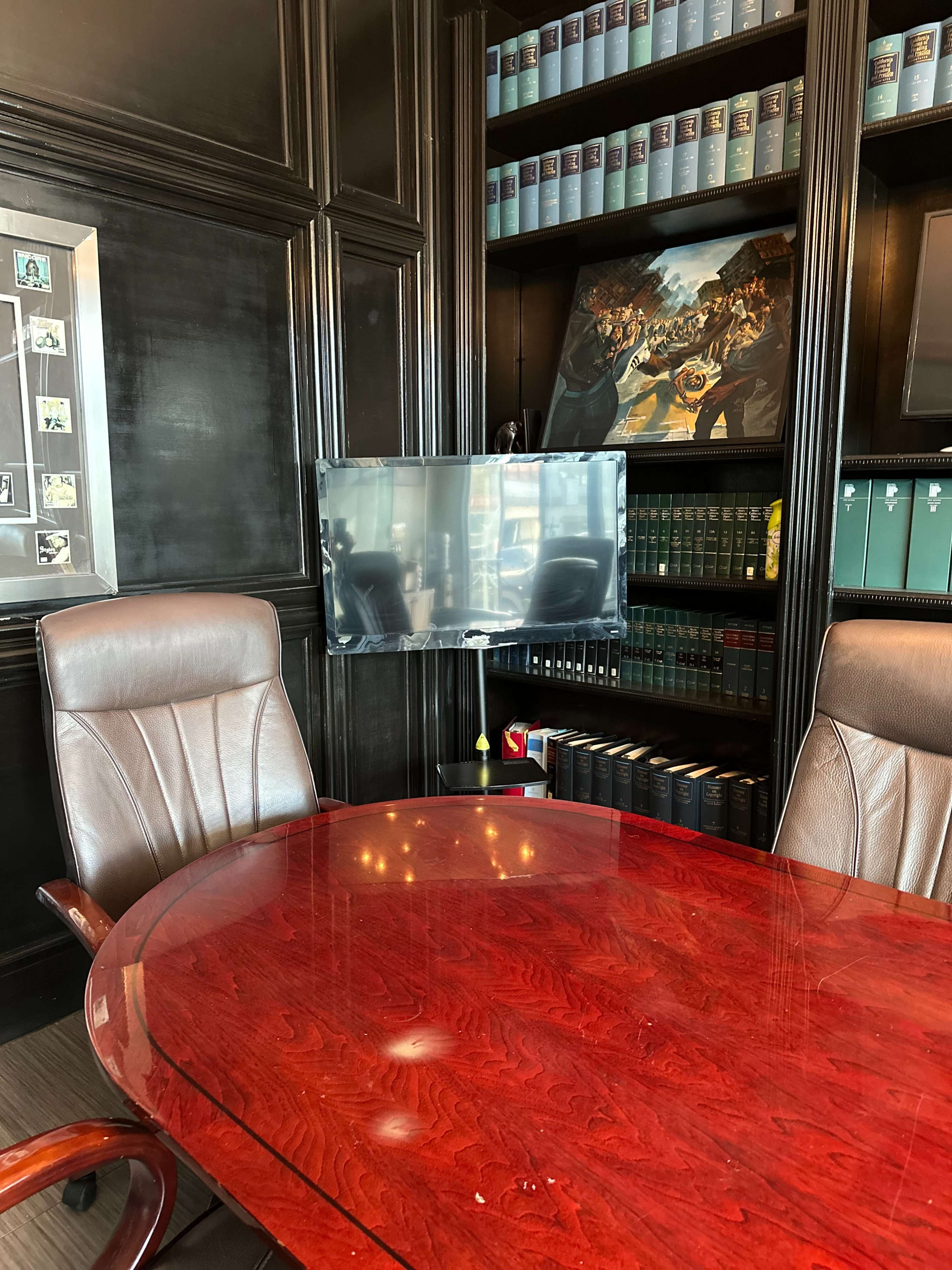 A polished wooden conference table is positioned in front of a wall-mounted TV, surrounded by leather chairs and a bookshelf filled with books and framed photos.