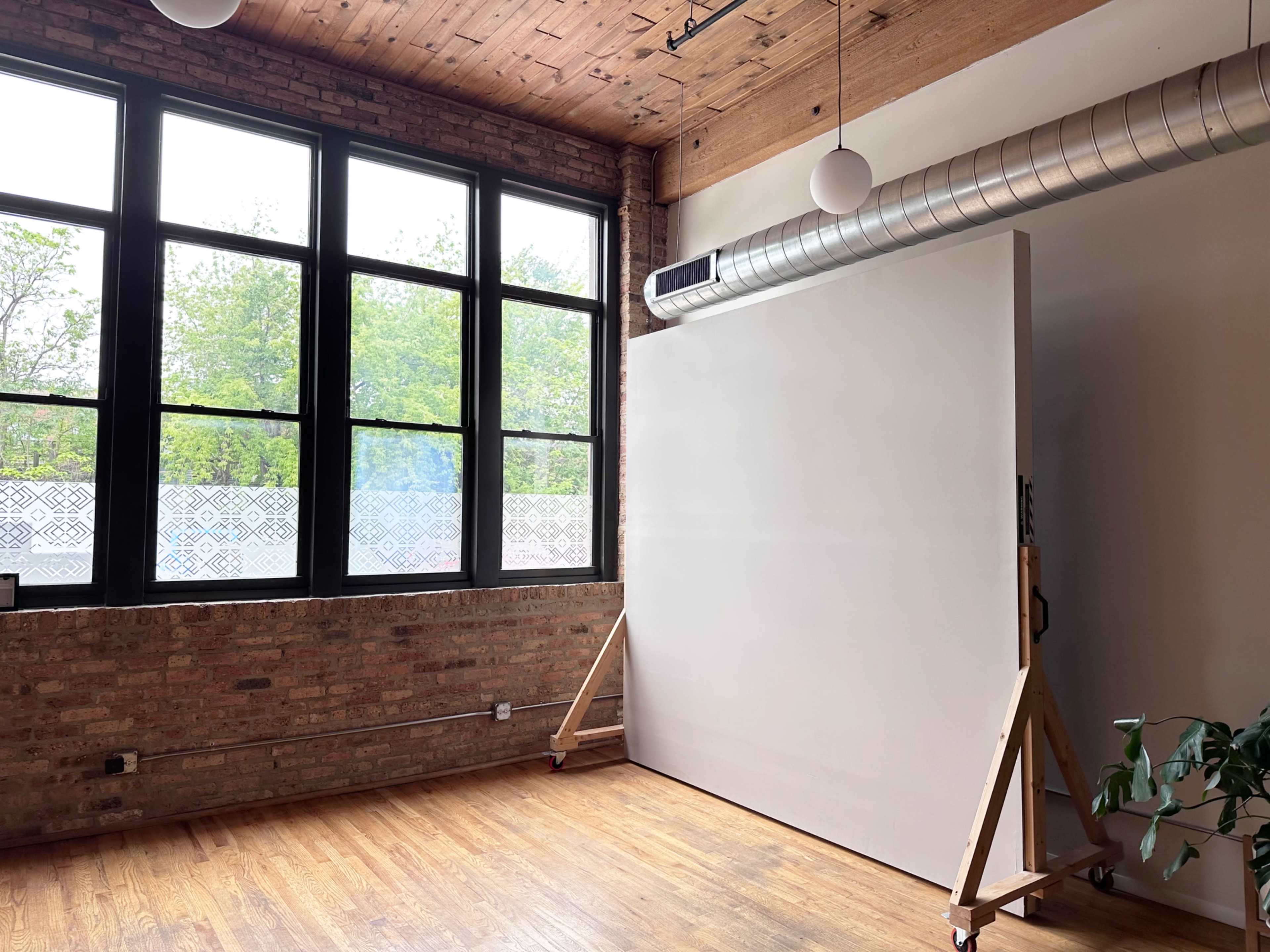 The image shows a spacious interior room with brick walls, large windows letting in natural light, a blank canvas on an easel, and a potted plant in the corner.