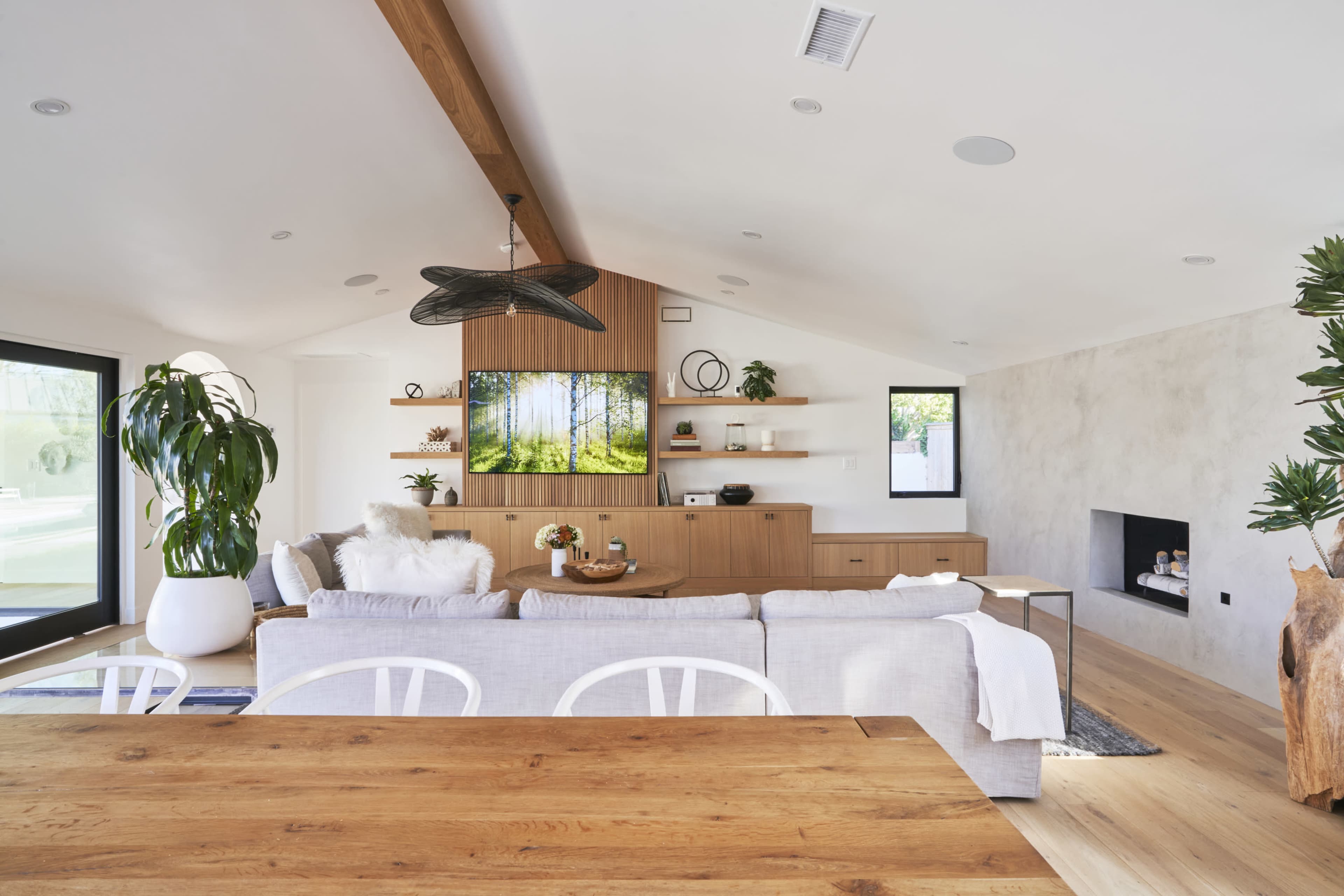 A modern, airy living room with a dining area featuring a wooden table, a light-colored sofa, a television mounted on a wooden wall unit, and large windows allowing natural light.