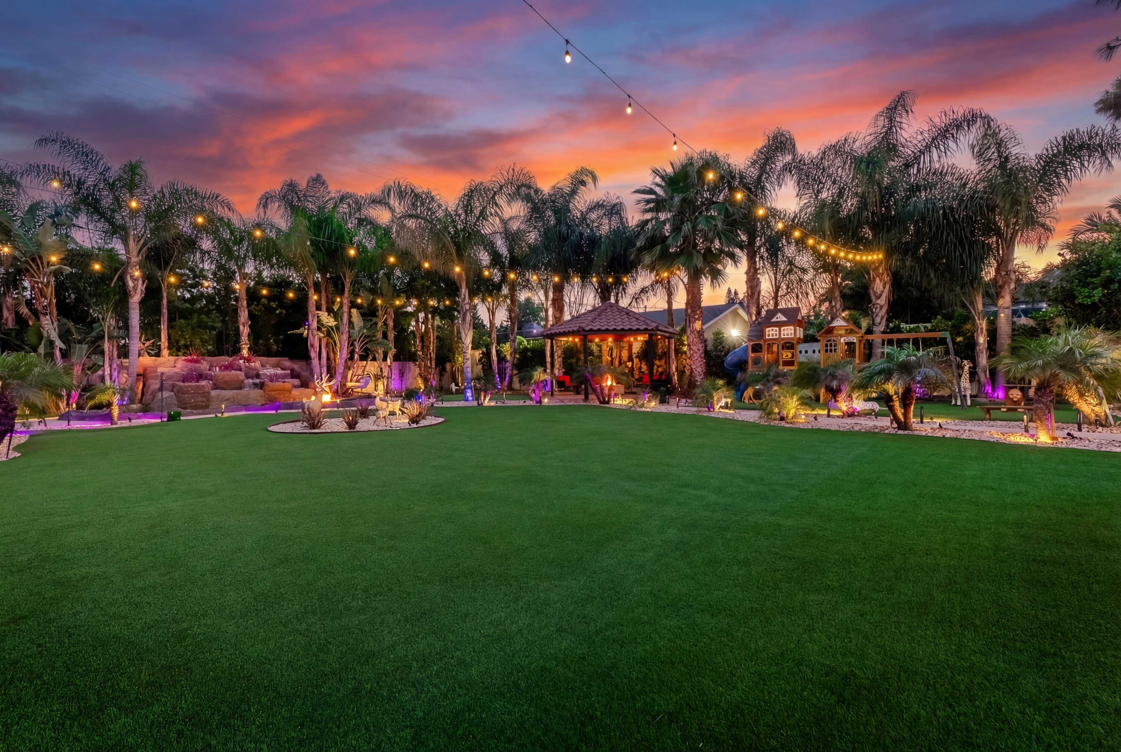 The image shows a well-lit backyard with palm trees, a landscaped lawn, and a gazebo under a colorful sunset sky.