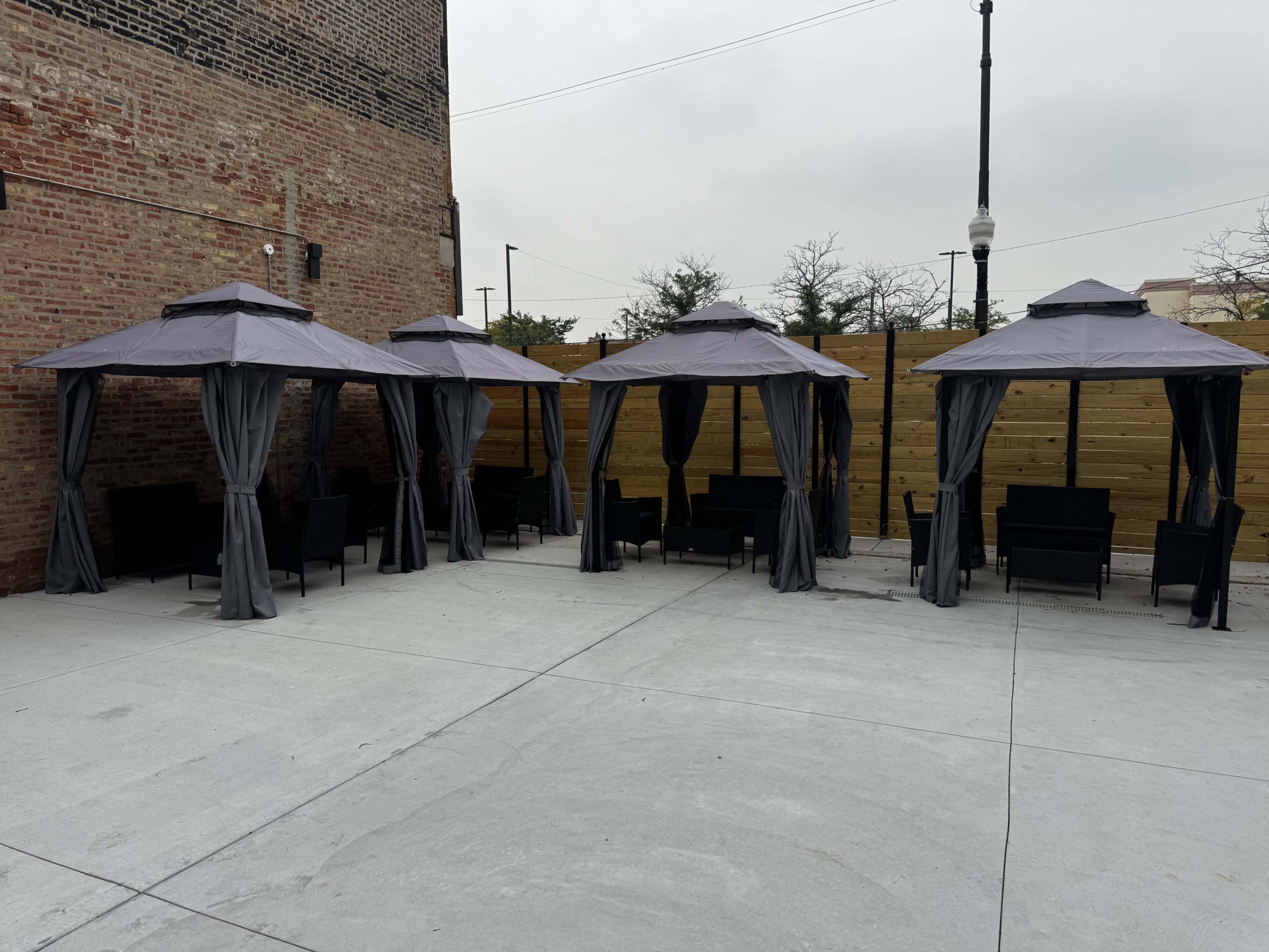 The image shows a series of four gray garden gazebos with curtains set up on a concrete patio, accompanied by black outdoor furniture.