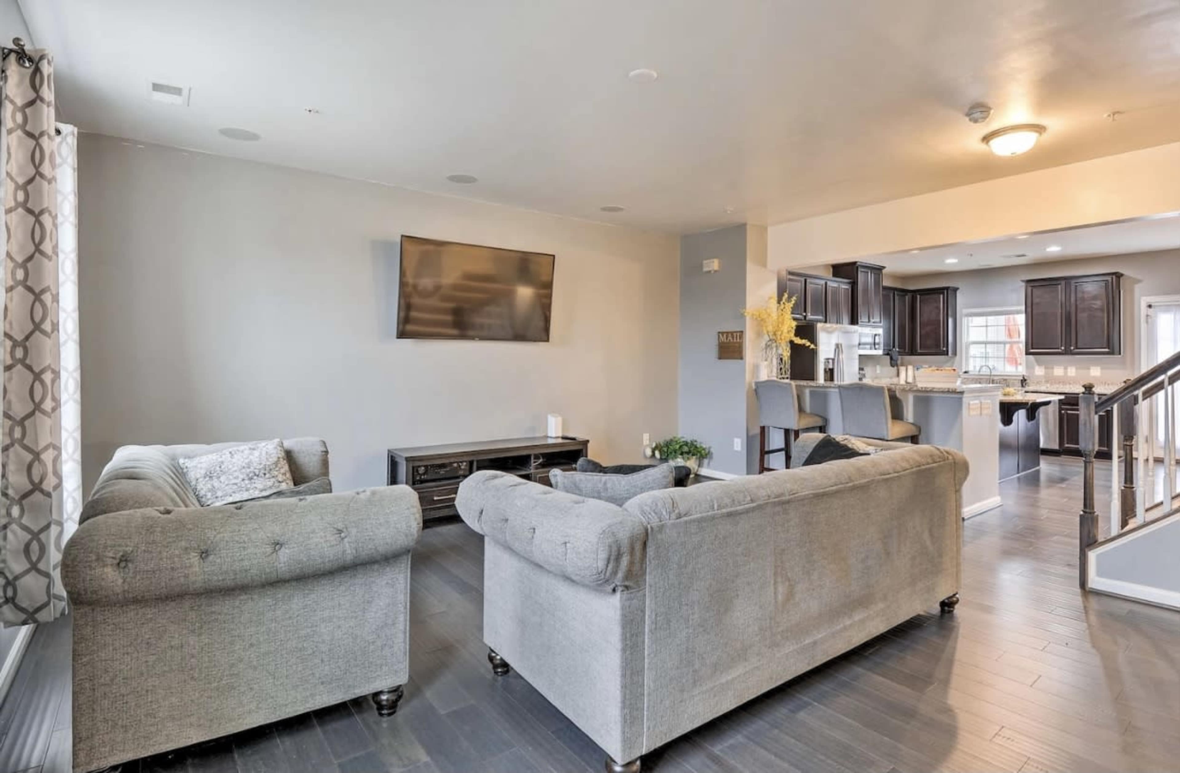 A modern living room with two gray couches facing a wall-mounted television, leading into a kitchen area with dark cabinetry.
