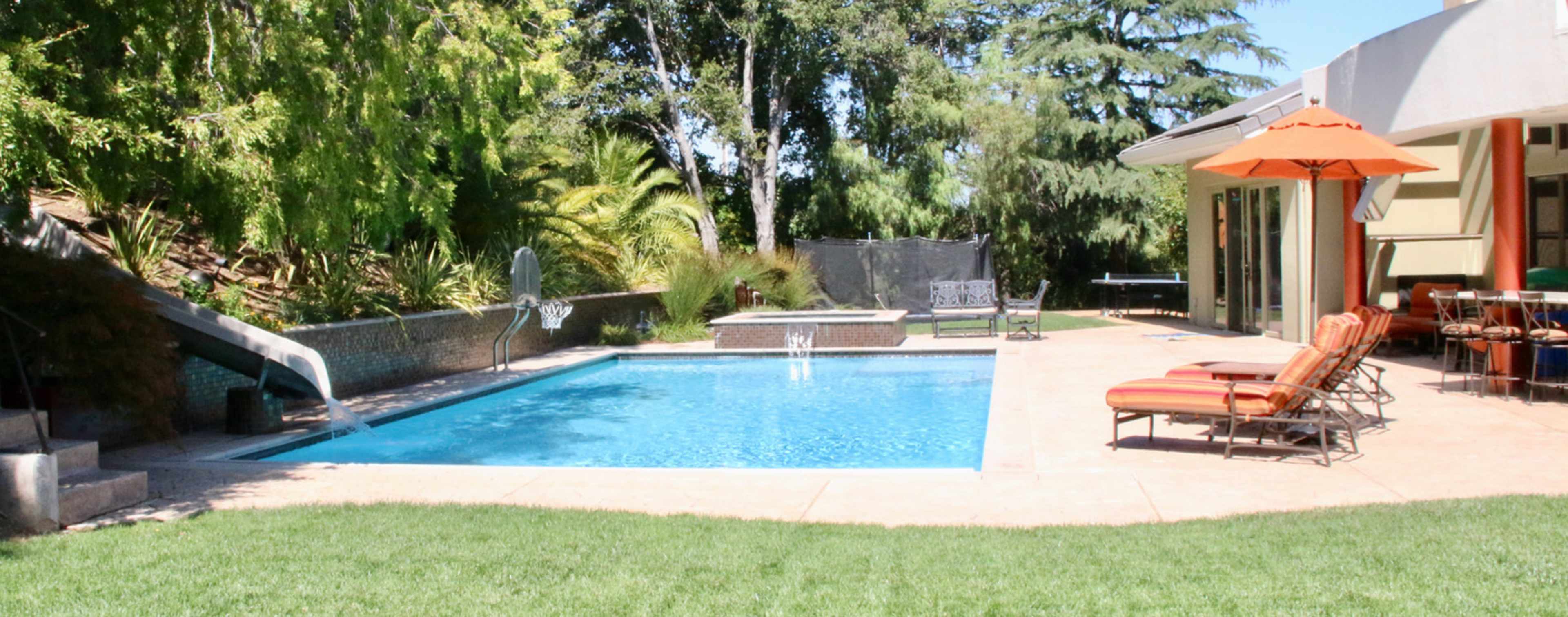 A backyard with a swimming pool surrounded by lounge chairs and an umbrella, with a lawn and trees in the background.
