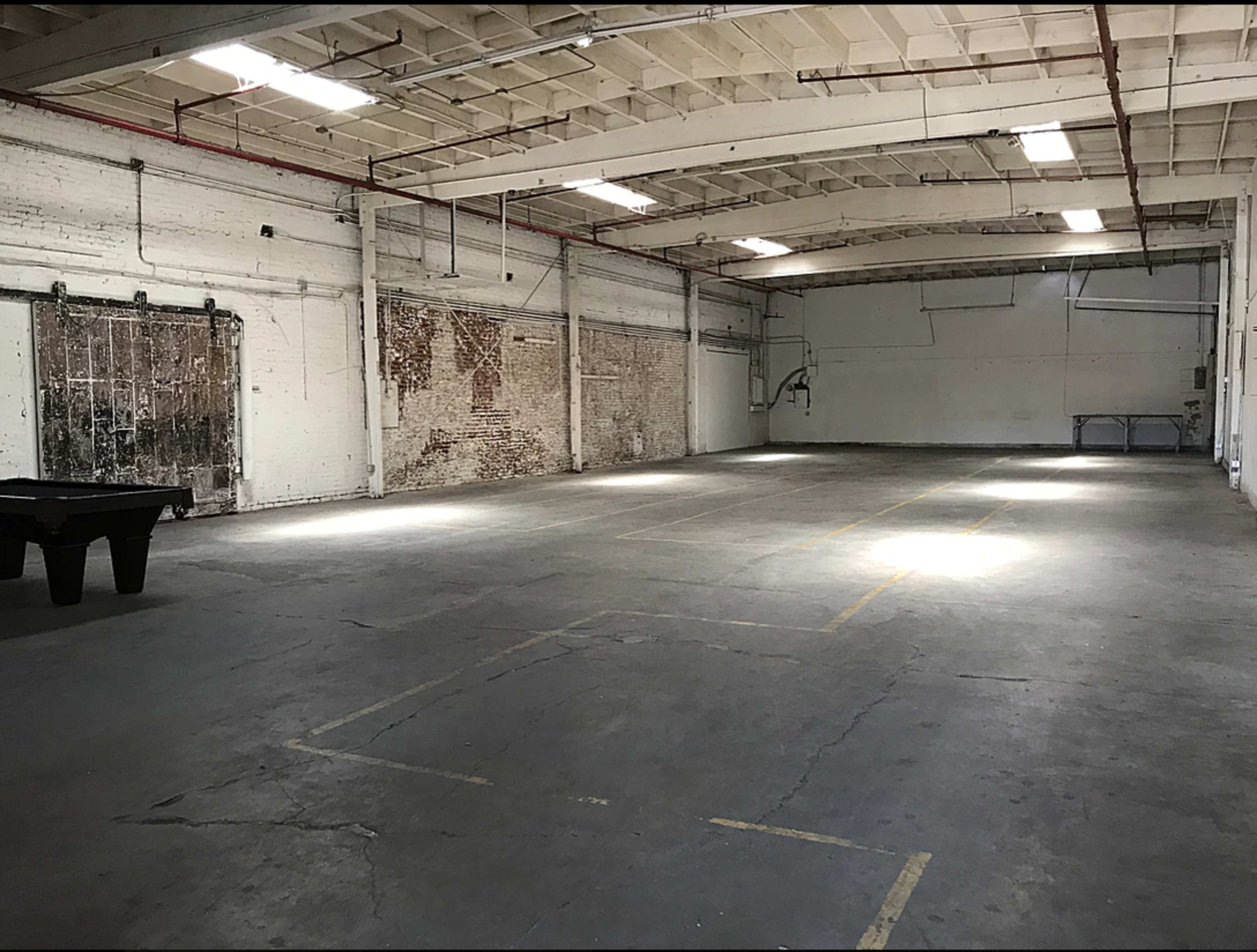 The image shows a large, empty industrial warehouse with concrete floors and exposed brick walls, featuring overhead lighting and minimal furnishings.