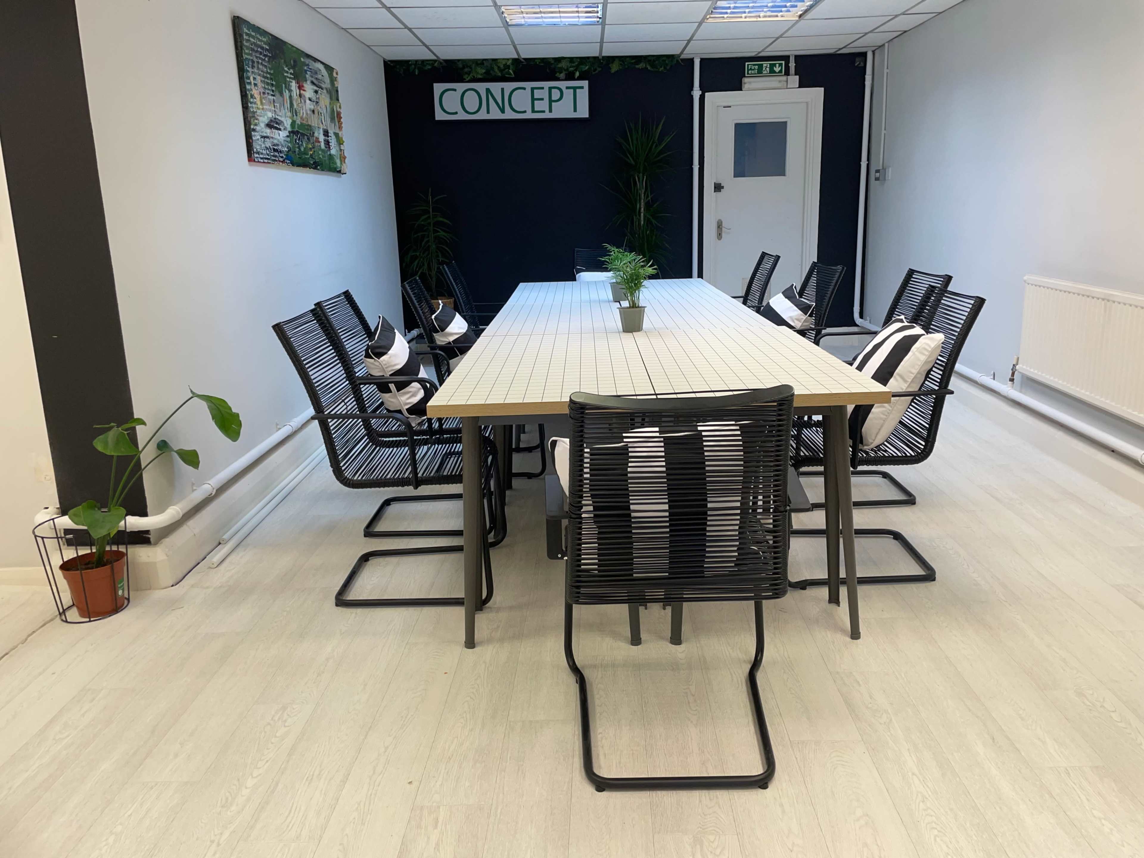 A long rectangular table surrounded by black chairs is set up in a bright, minimalist meeting room with green plants and a sign that reads "CONCEPT."