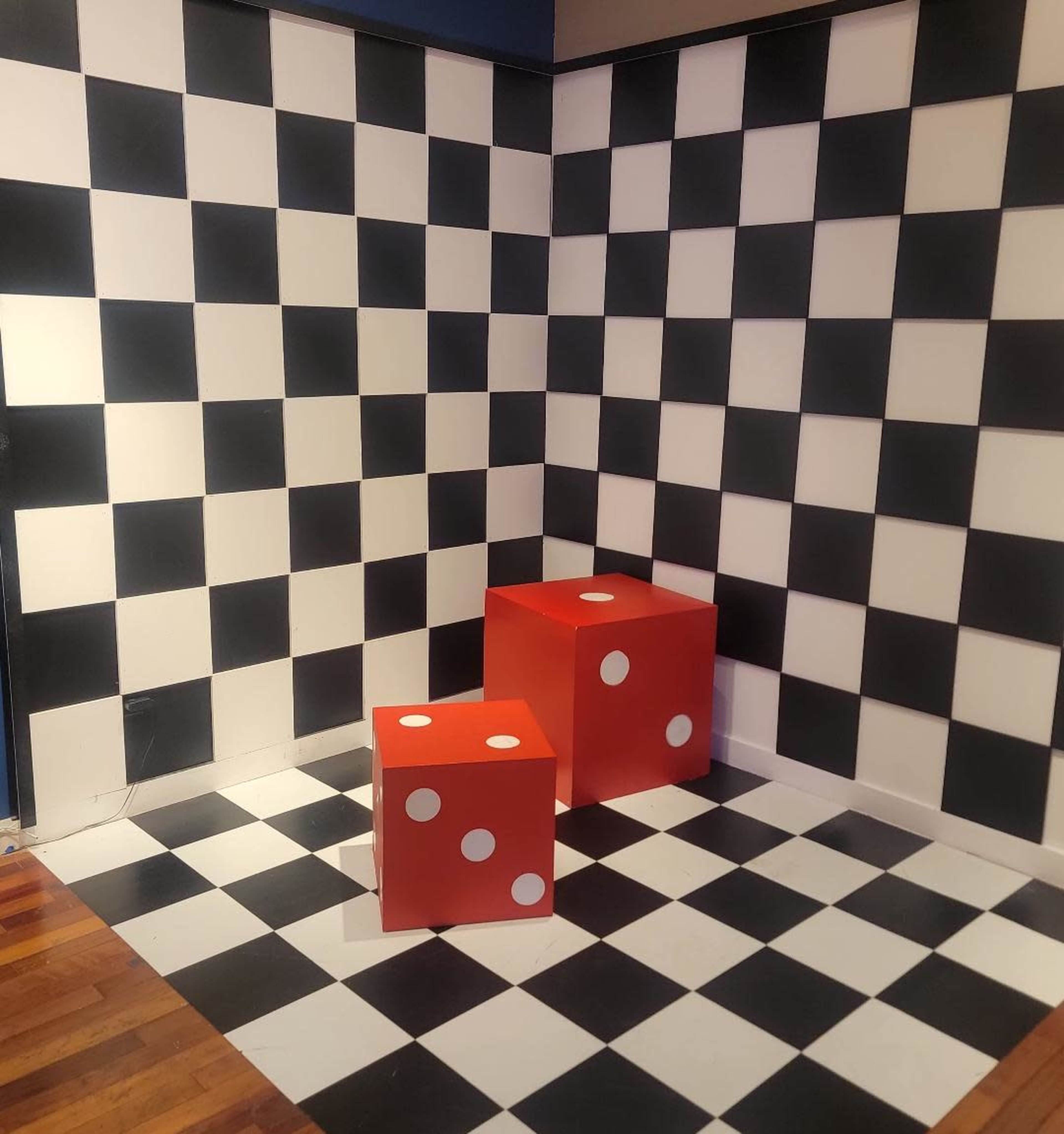 A corner room with black and white checkered walls and floor, featuring two large red dice with white dots.