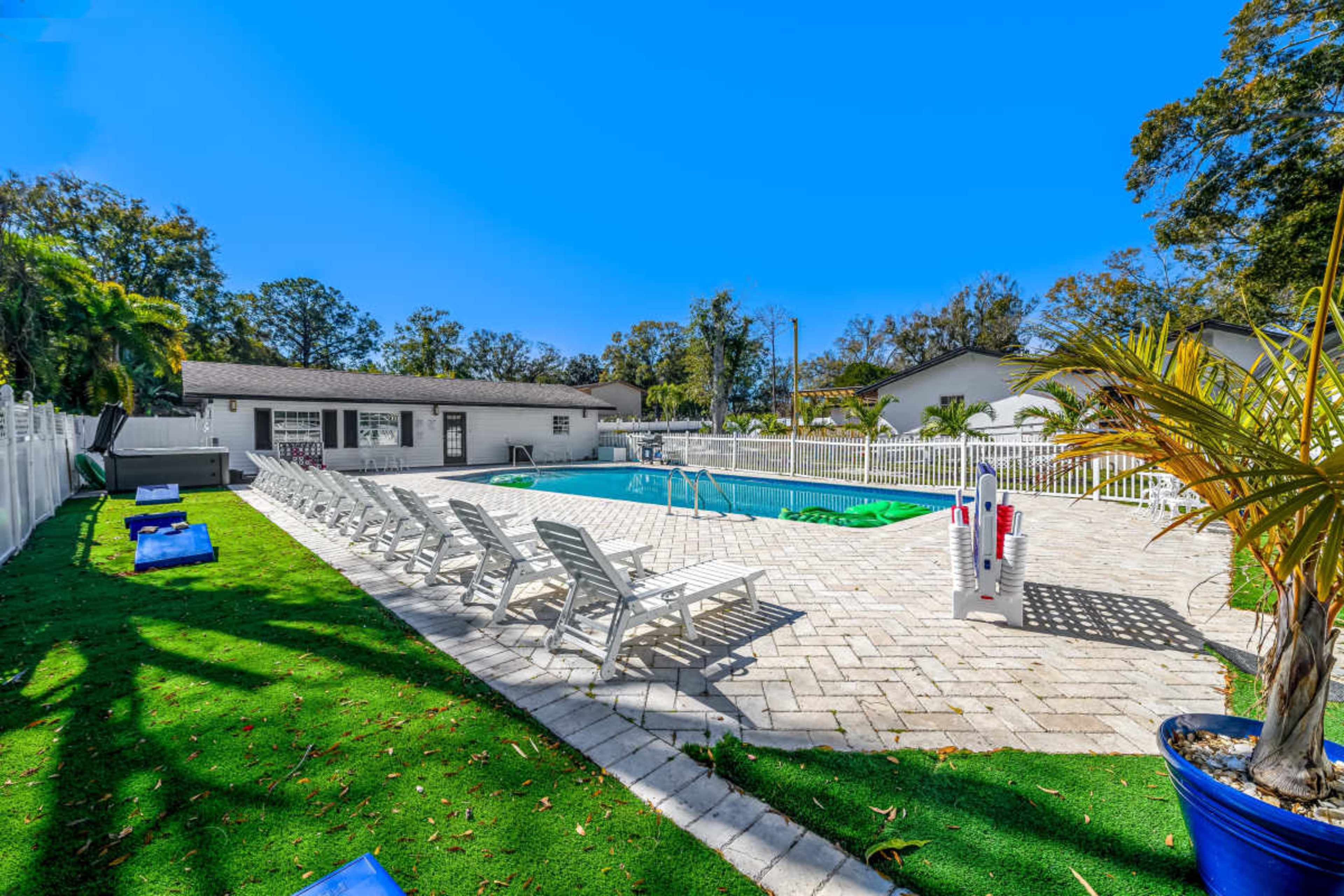 The image shows a swimming pool area surrounded by lounge chairs, with a grassy section and palm trees, set against a clear blue sky.