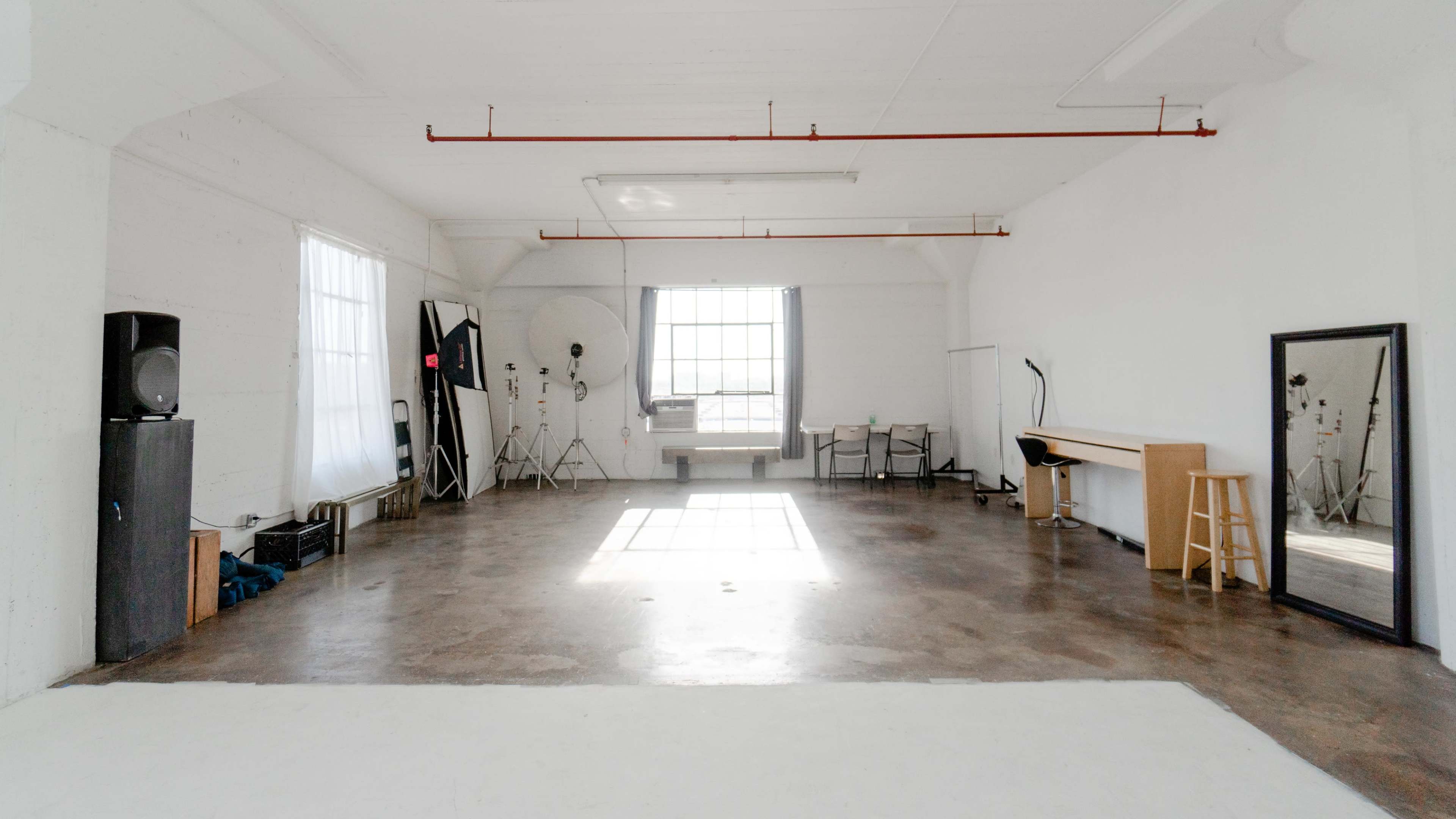 A spacious, empty room with white walls, large windows letting in natural light, and minimal furniture, including a table, stool, and mirror.
