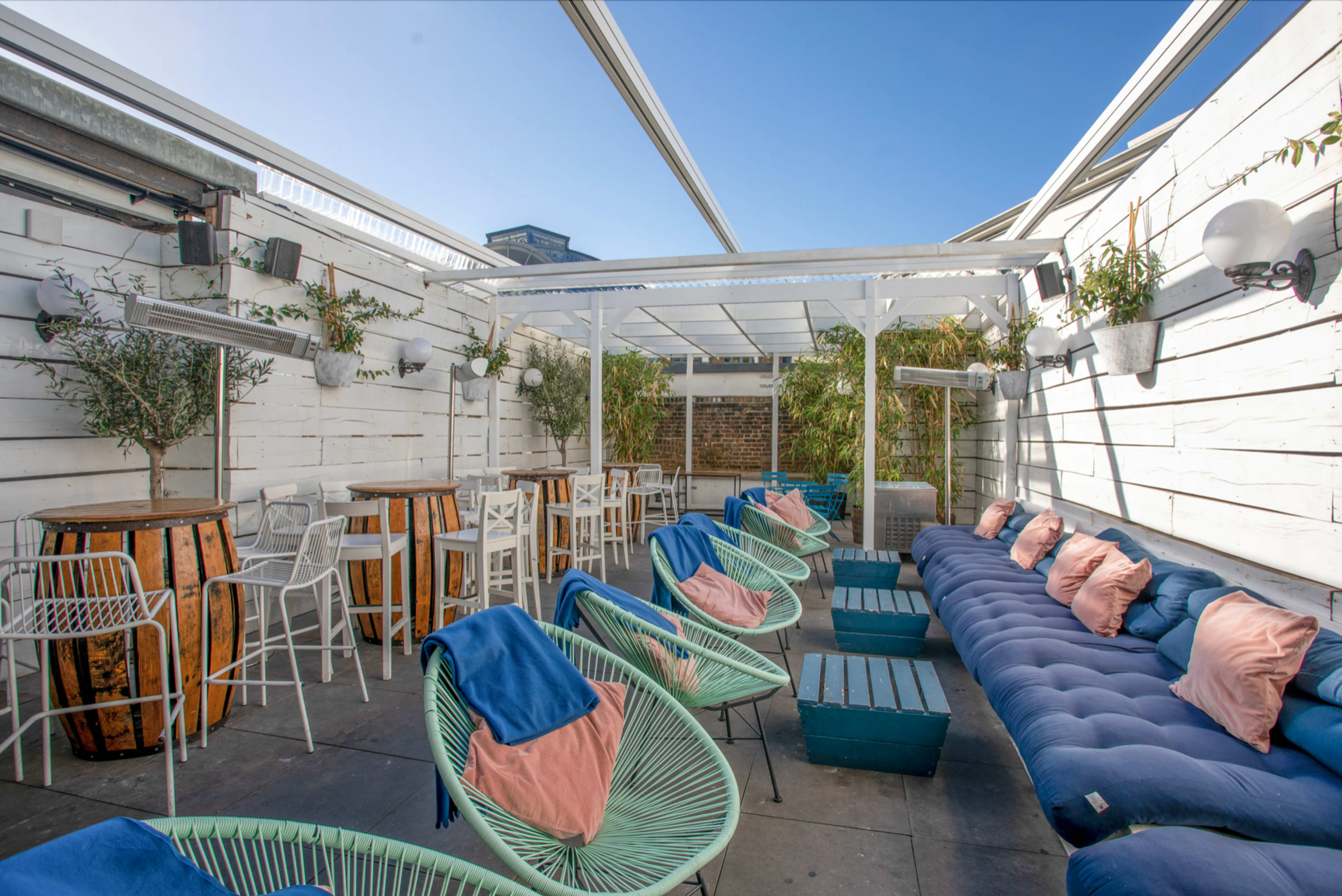 A rooftop lounge area with white walls, a mix of seating options including green chairs and a blue sofa, and wooden tables under a glass canopy.