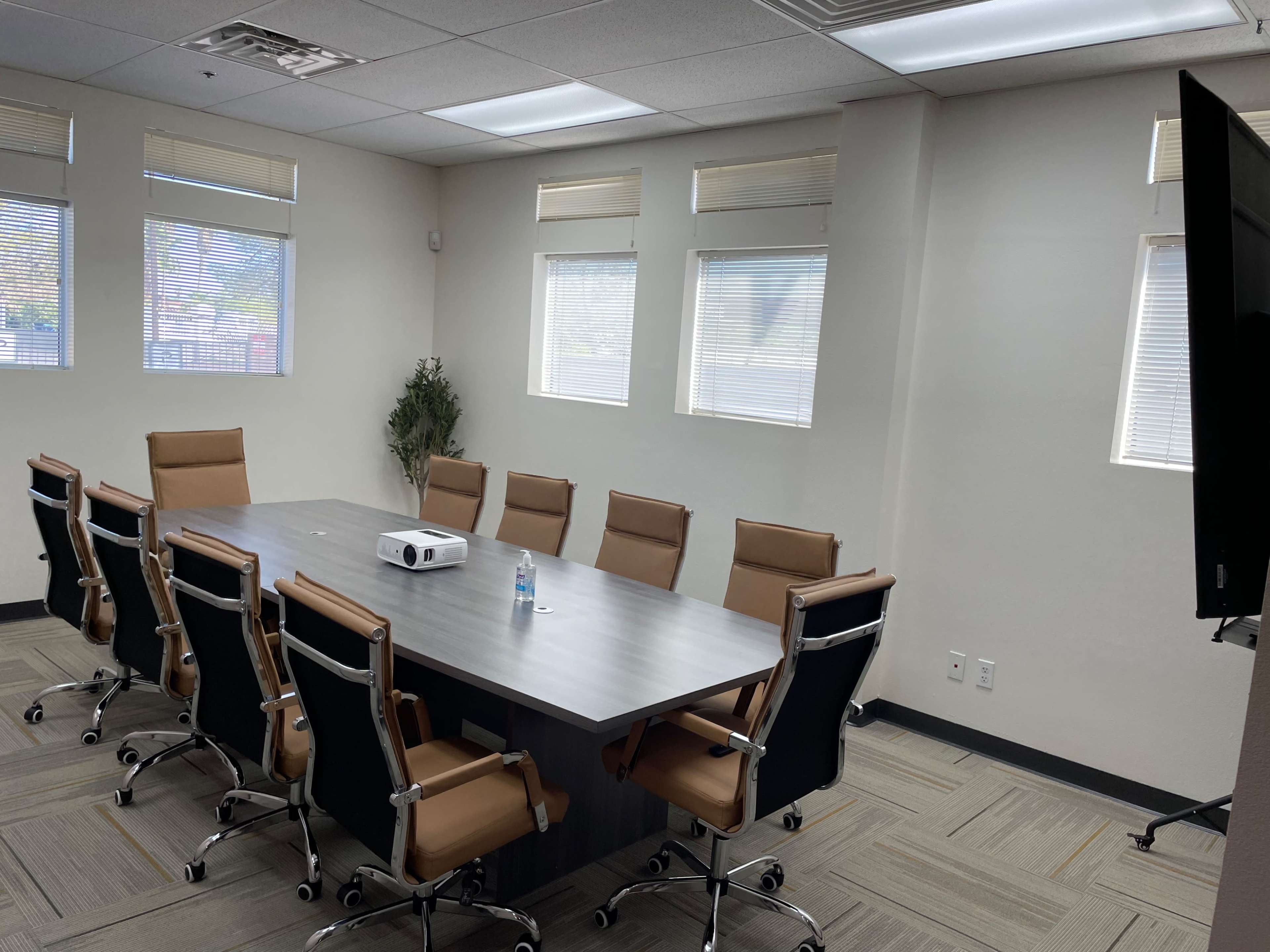 A conference room with a long rectangular table surrounded by eight brown chairs, a projector on the table, and large windows allowing natural light.