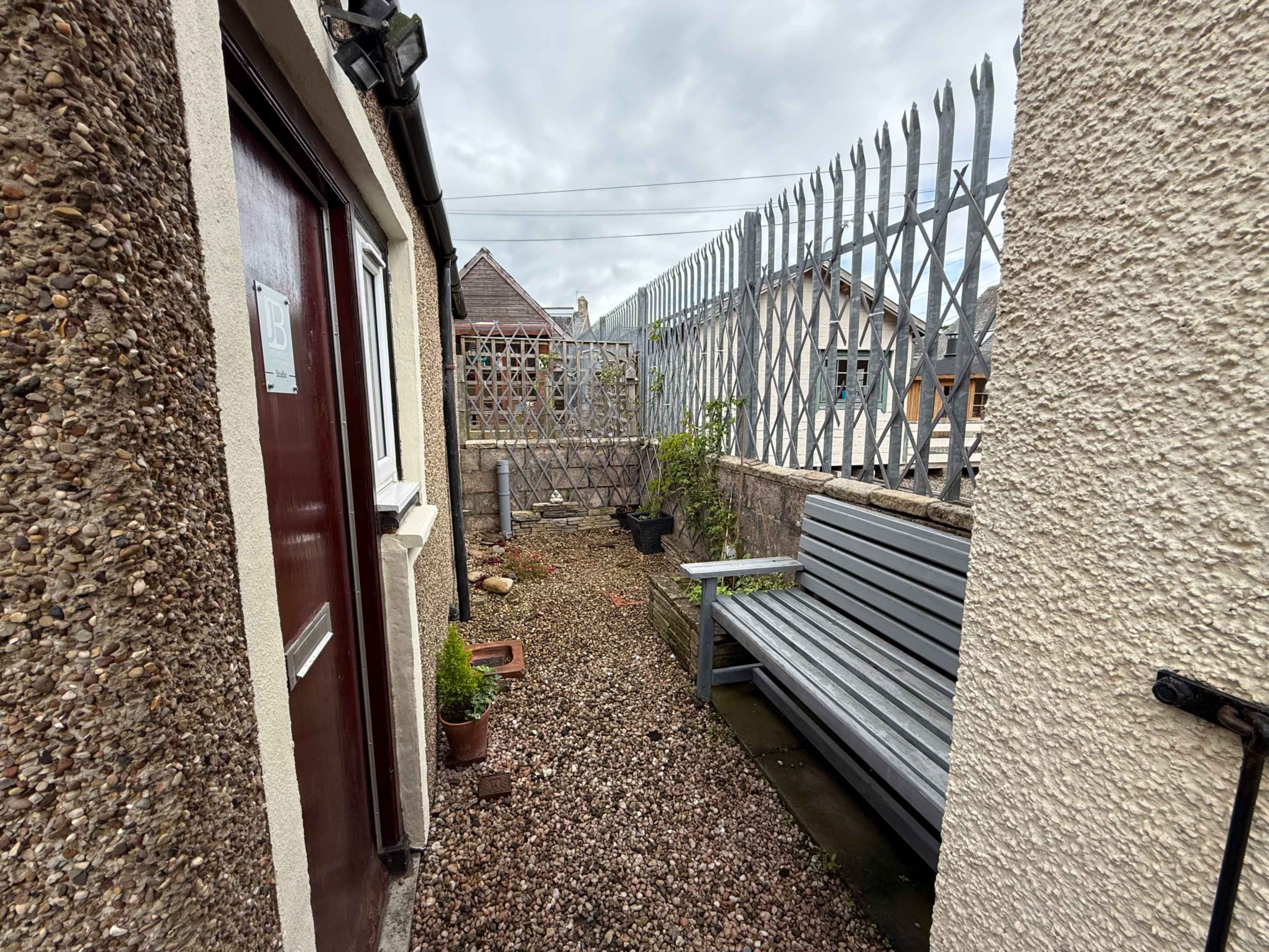 The image shows a narrow outdoor space with a gravel path, a wooden bench, potted plants, and a fence in the background.