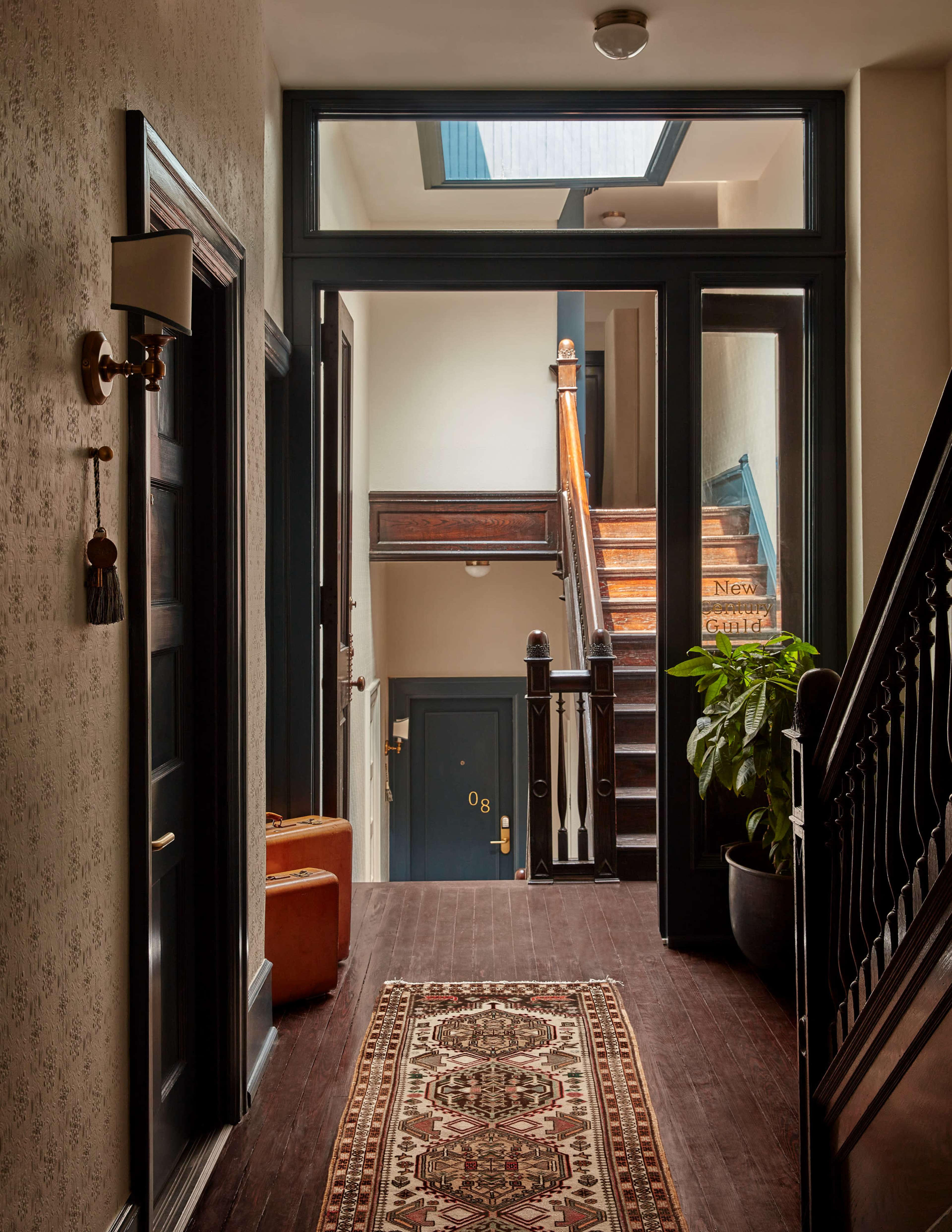 A well-lit hallway features a staircase, decorative wallpaper, and a potted plant beside a patterned rug.