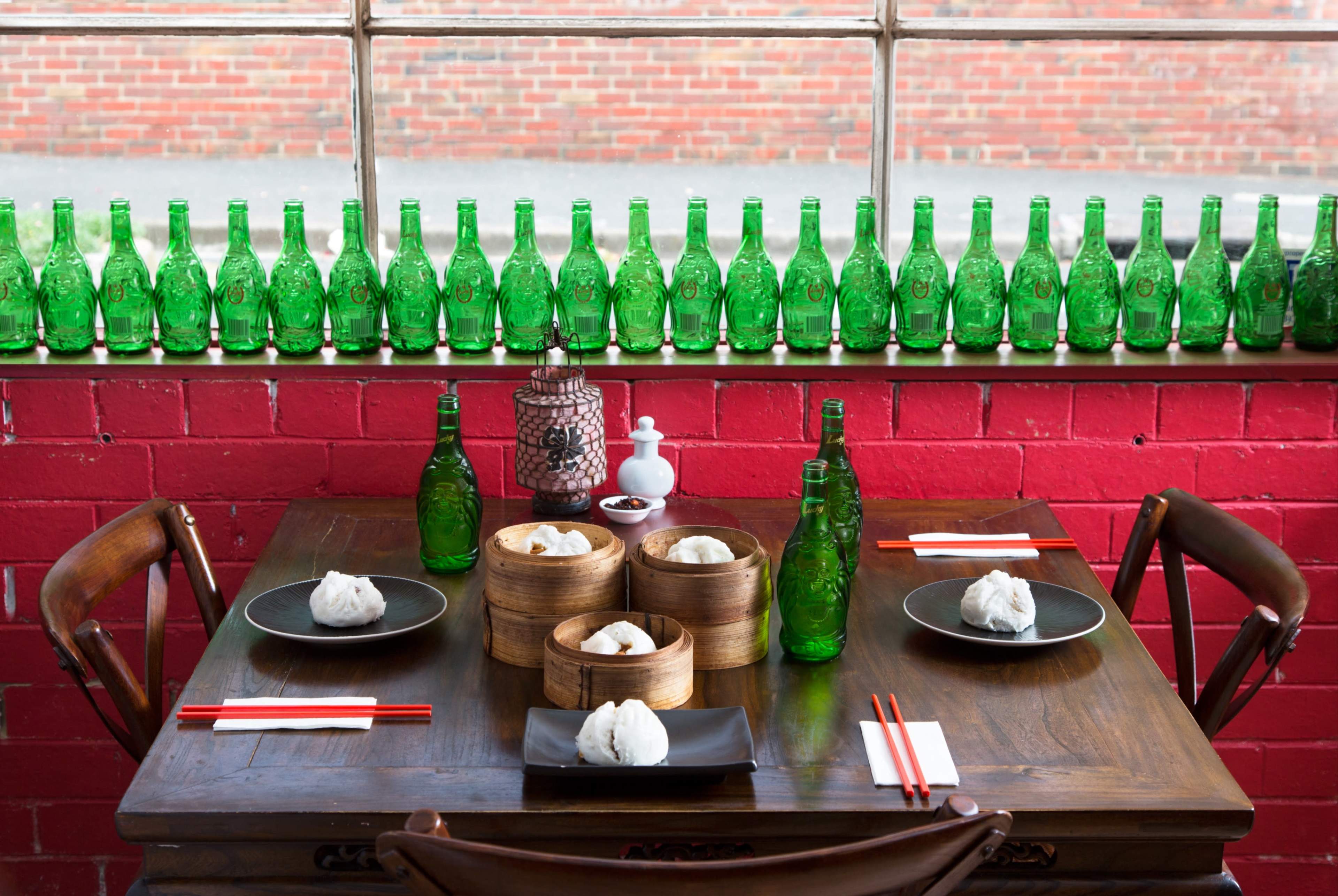 A wooden table is set with bamboo steamers containing buns, surrounded by green bottles and chopsticks, against a backdrop of red bricks and a window.