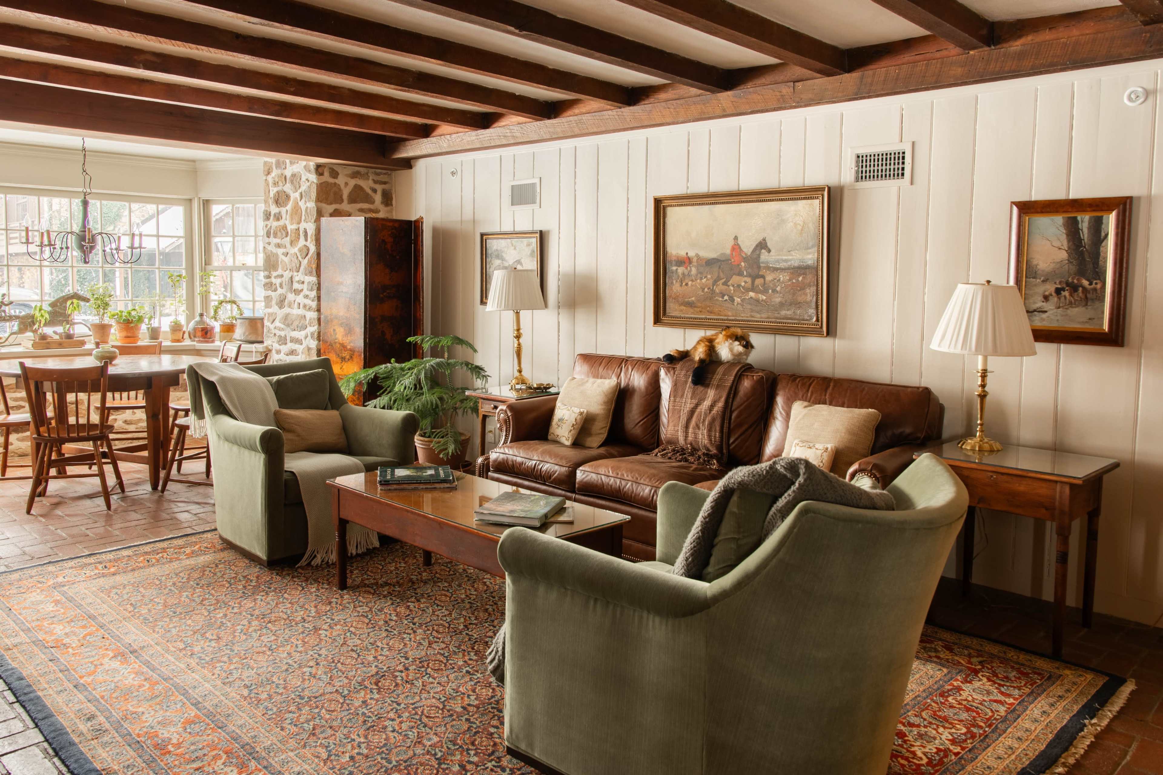 The image shows a cozy living room with a leather sofa, two green chairs, a wooden coffee table, and decorative paintings on the walls, featuring exposed beams and a nearby dining area.