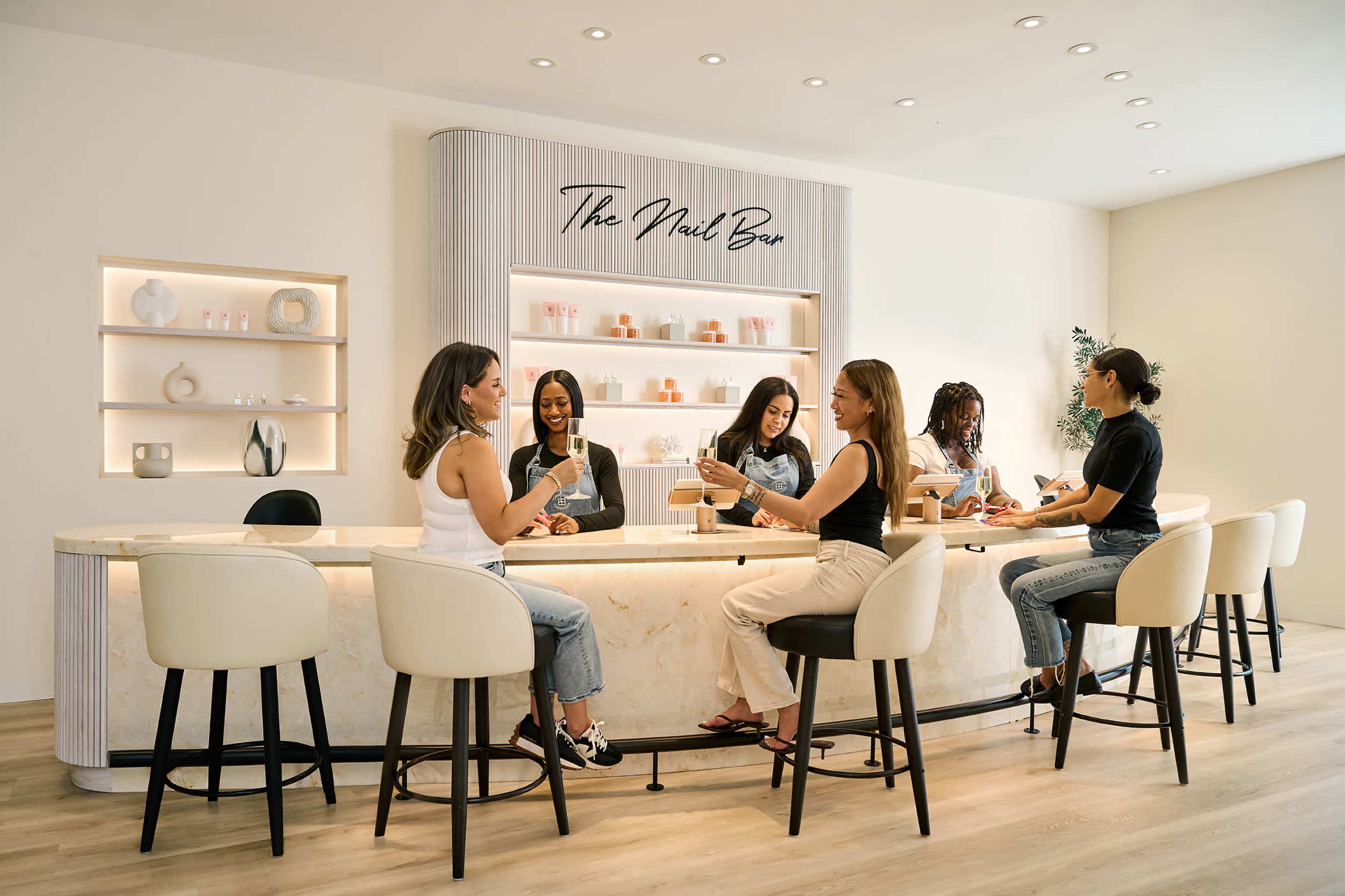 A stylish nail salon features a spacious bar where several women are seated, enjoying drinks and socializing while receiving nail services.