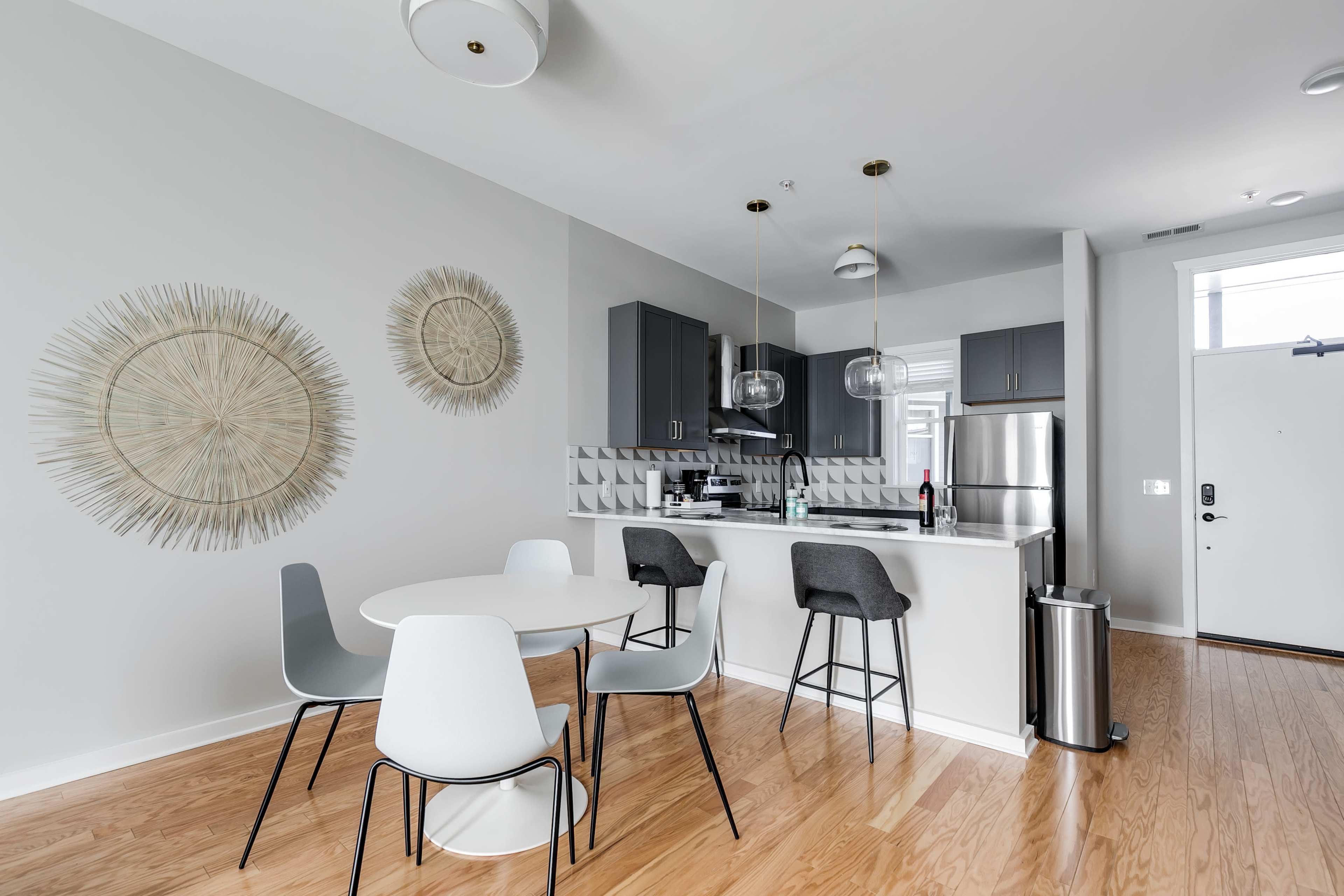 A modern kitchen and dining area features a round table surrounded by chairs, with light-colored cabinetry and decorative wall elements.