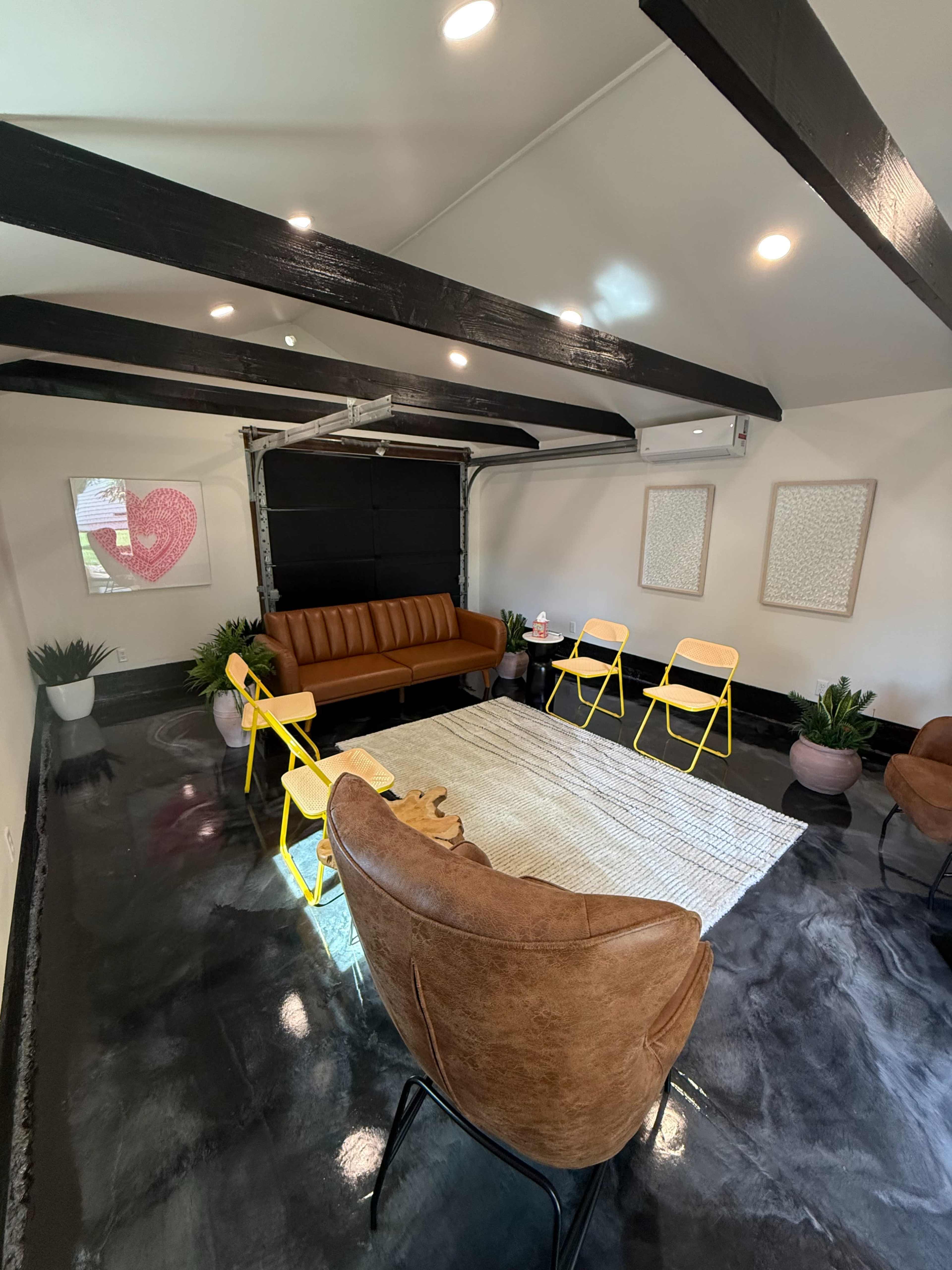 The image shows a modern interior space with a brown sofa, yellow chairs, a round rug, and plants in pots, featuring exposed beams on the ceiling.