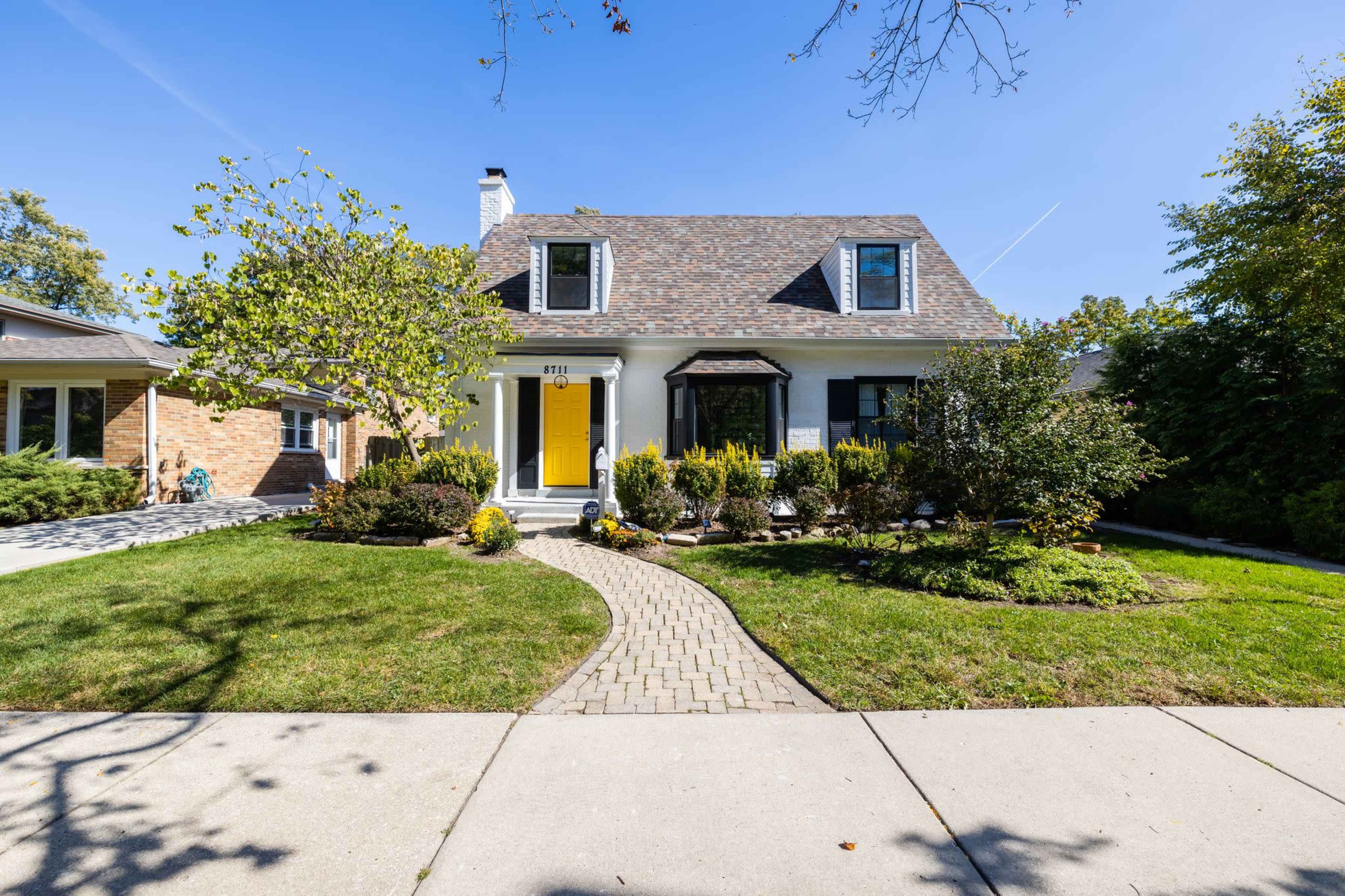 A two-story house with a yellow front door and a landscaped yard features a walkway made of bricks leading to the entrance.