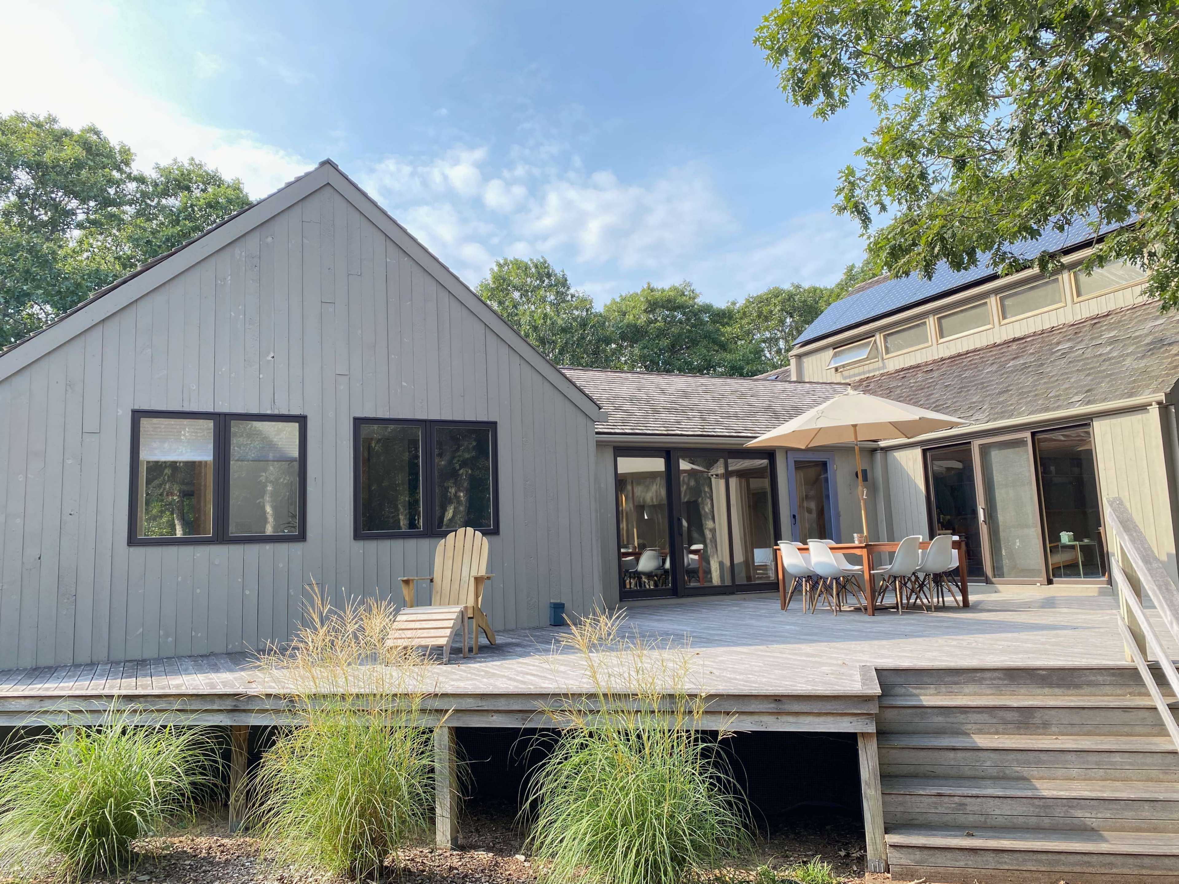 A modern wood-sided house features a spacious deck with outdoor seating and an umbrella.