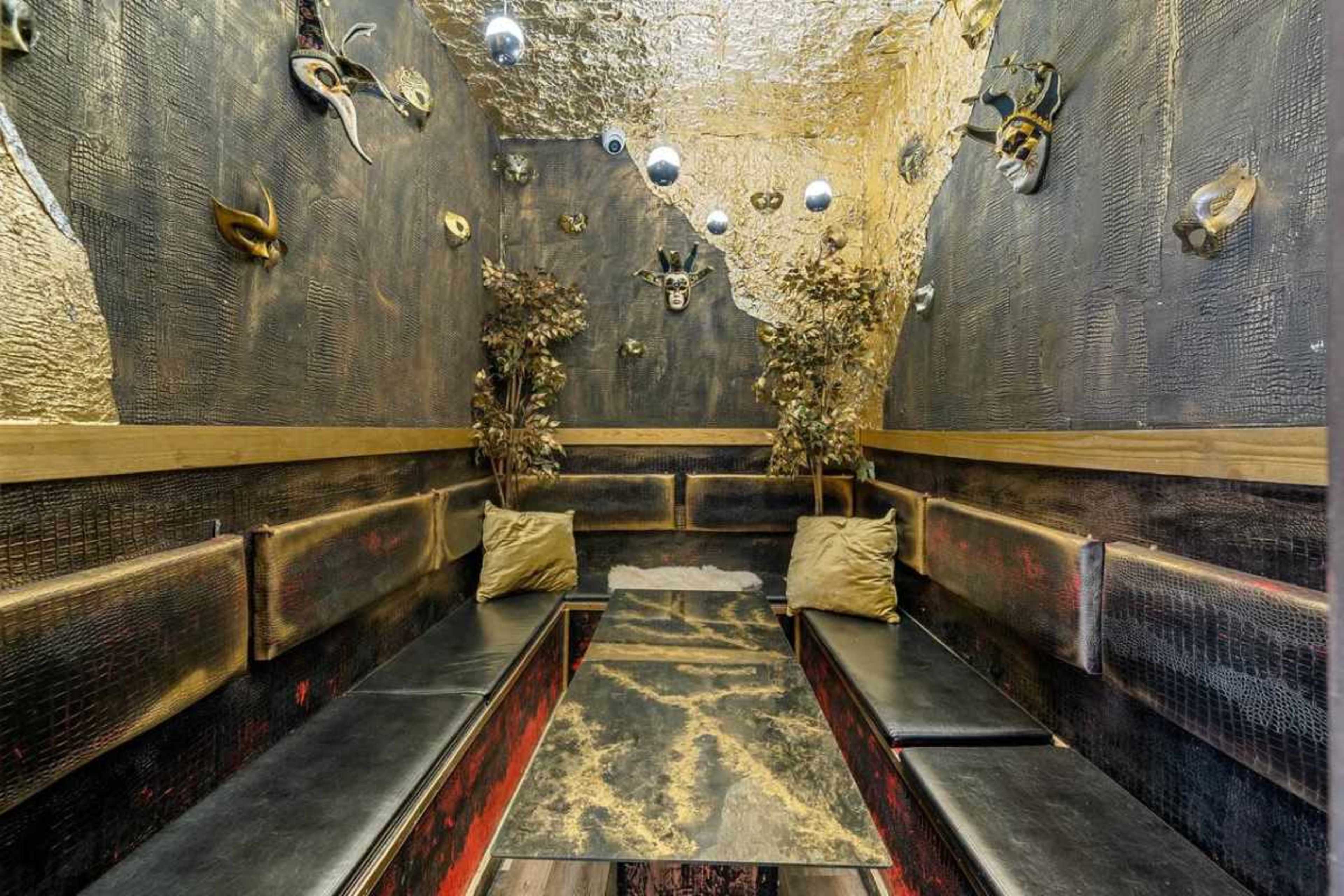 The image shows a dimly lit, decorative lounge area with black walls, a metallic ceiling, and a central table surrounded by seating.