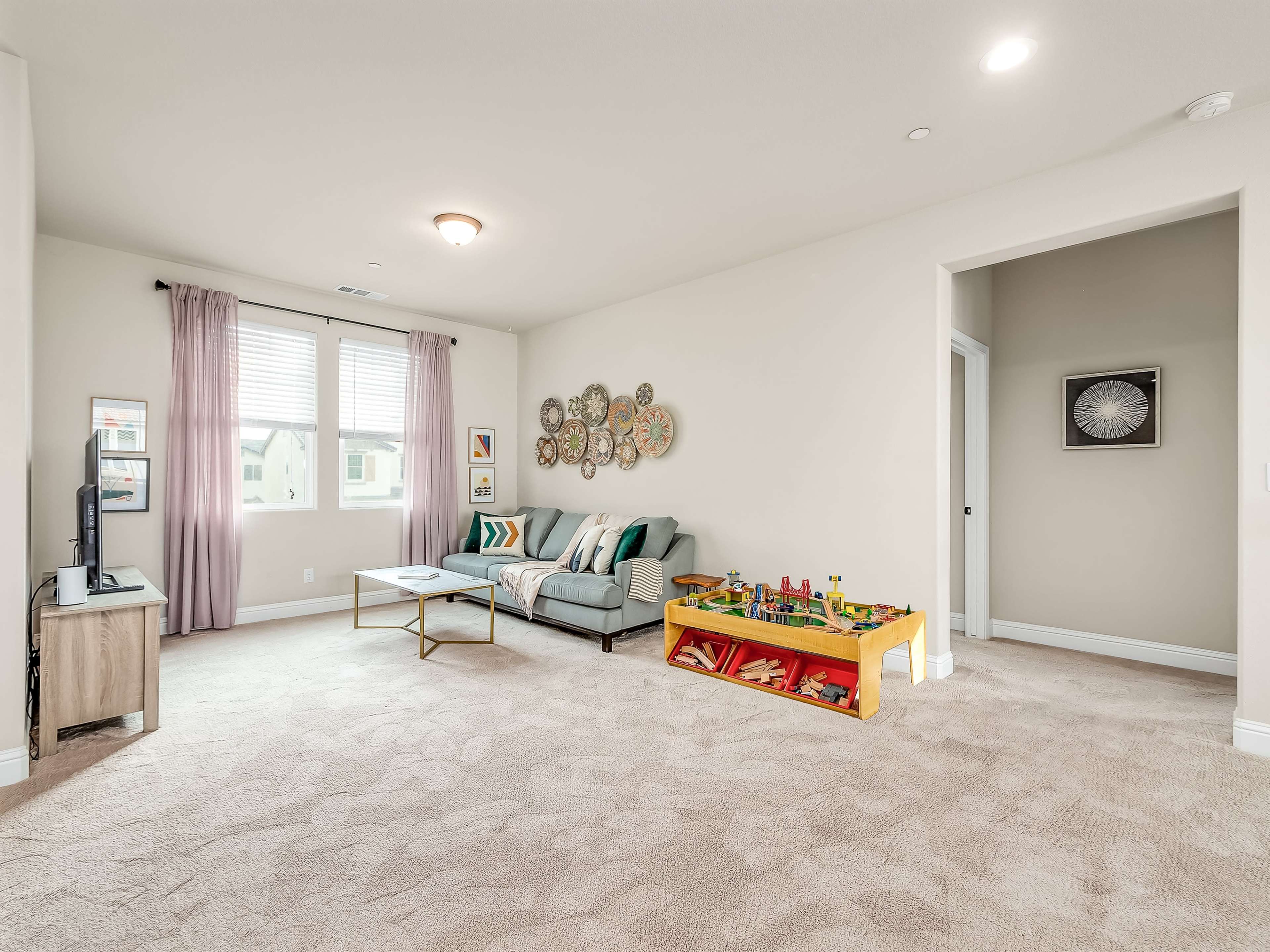 A furnished living room features a gray sofa, a coffee table, and a play area with toys, illuminated by natural light from two windows.