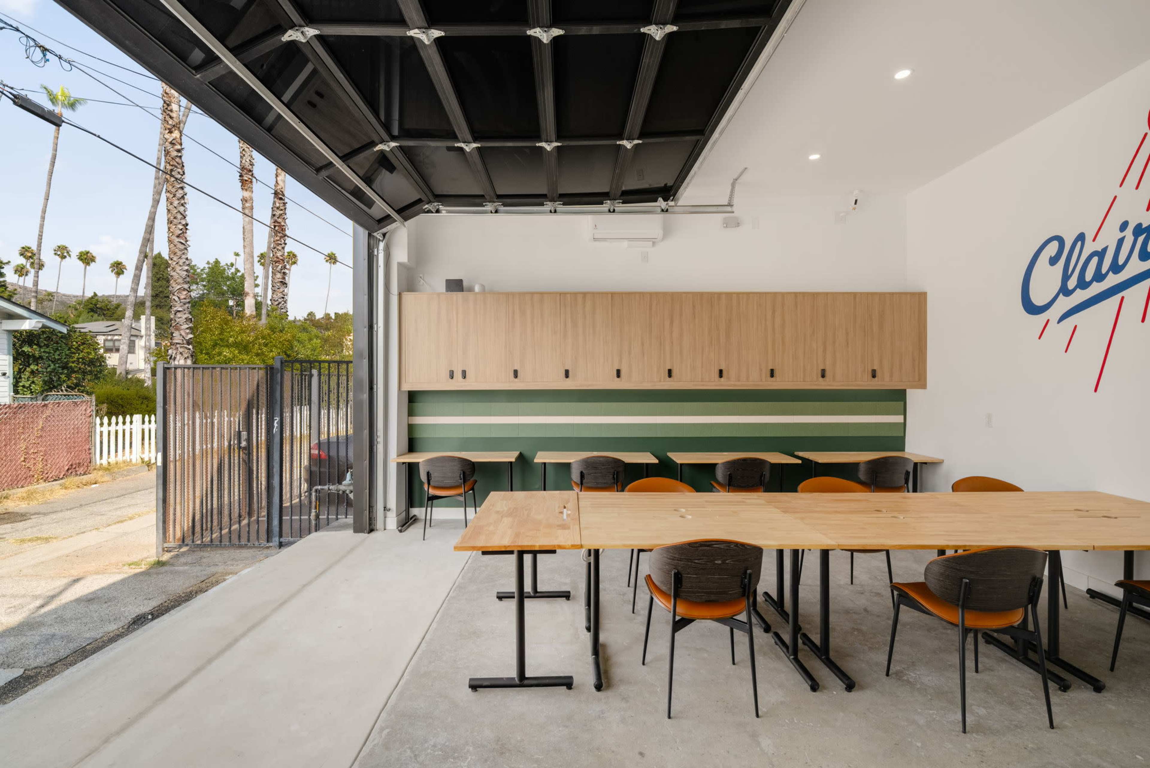 The image shows a modern, open space with a large garage door, several wooden tables, and chairs, along with storage cabinets on the wall decorated with a logo.