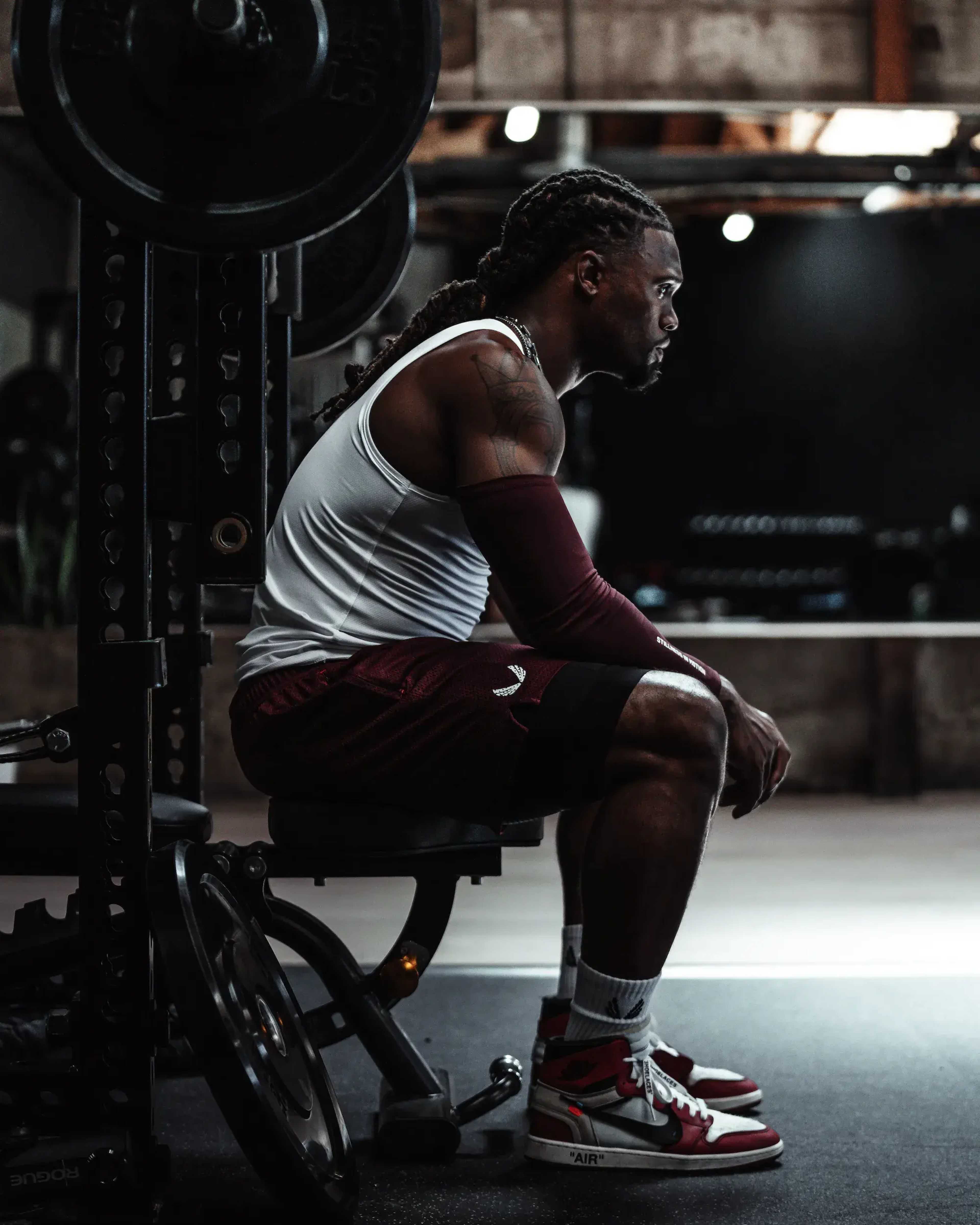 A muscular man sits on a weight bench in a gym, wearing a tank top and shorts, with weightlifting equipment in the background.