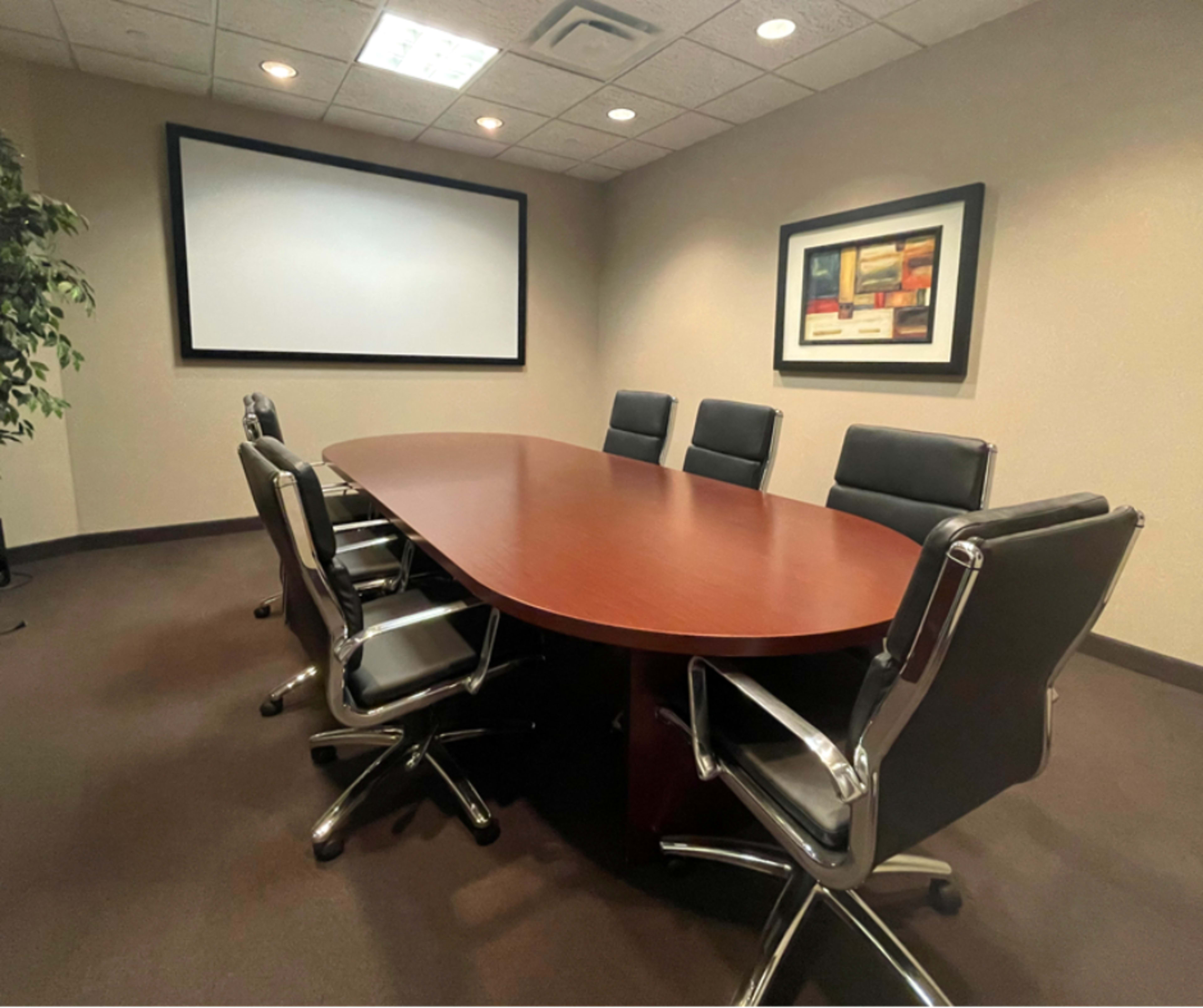 The image shows a conference room with a large oval wooden table surrounded by black leather chairs and a projection screen on the wall.