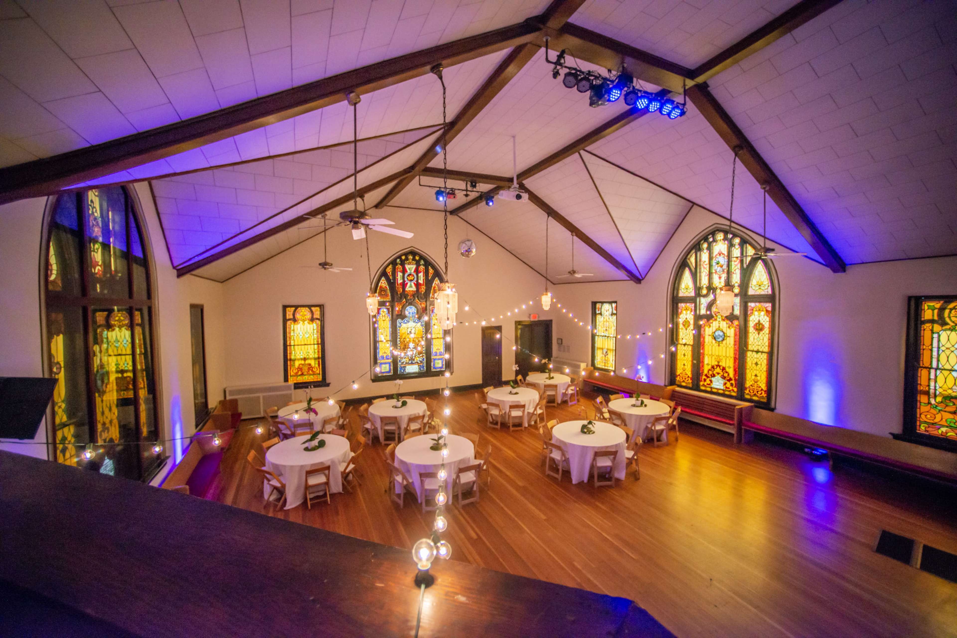 The interior of a hall features round tables arranged on a wooden floor beneath stained glass windows, with hanging lights and a wood-beamed ceiling.
