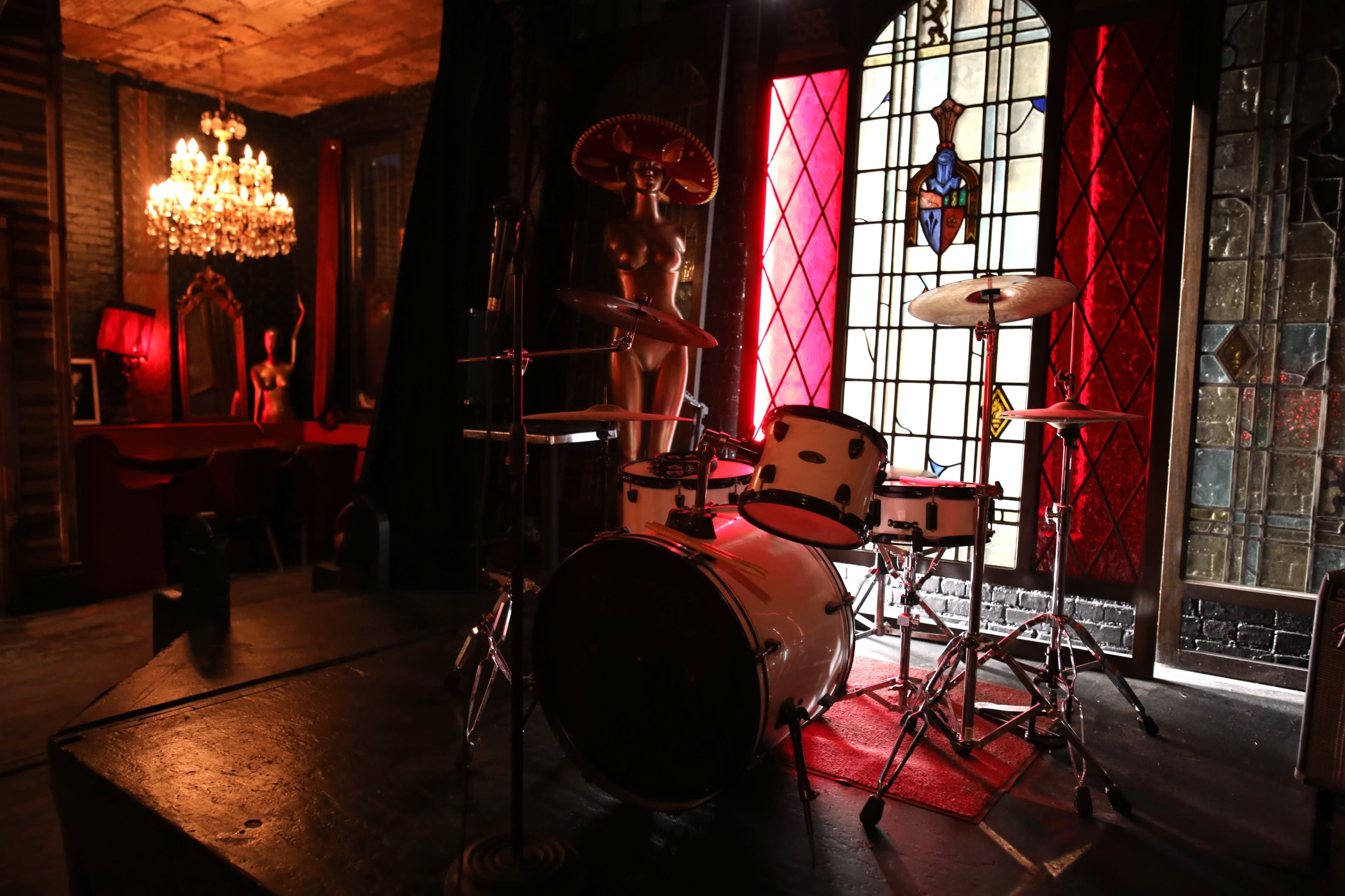 A drum set is positioned on a stage in a dimly lit room with stained glass windows and a chandelier.