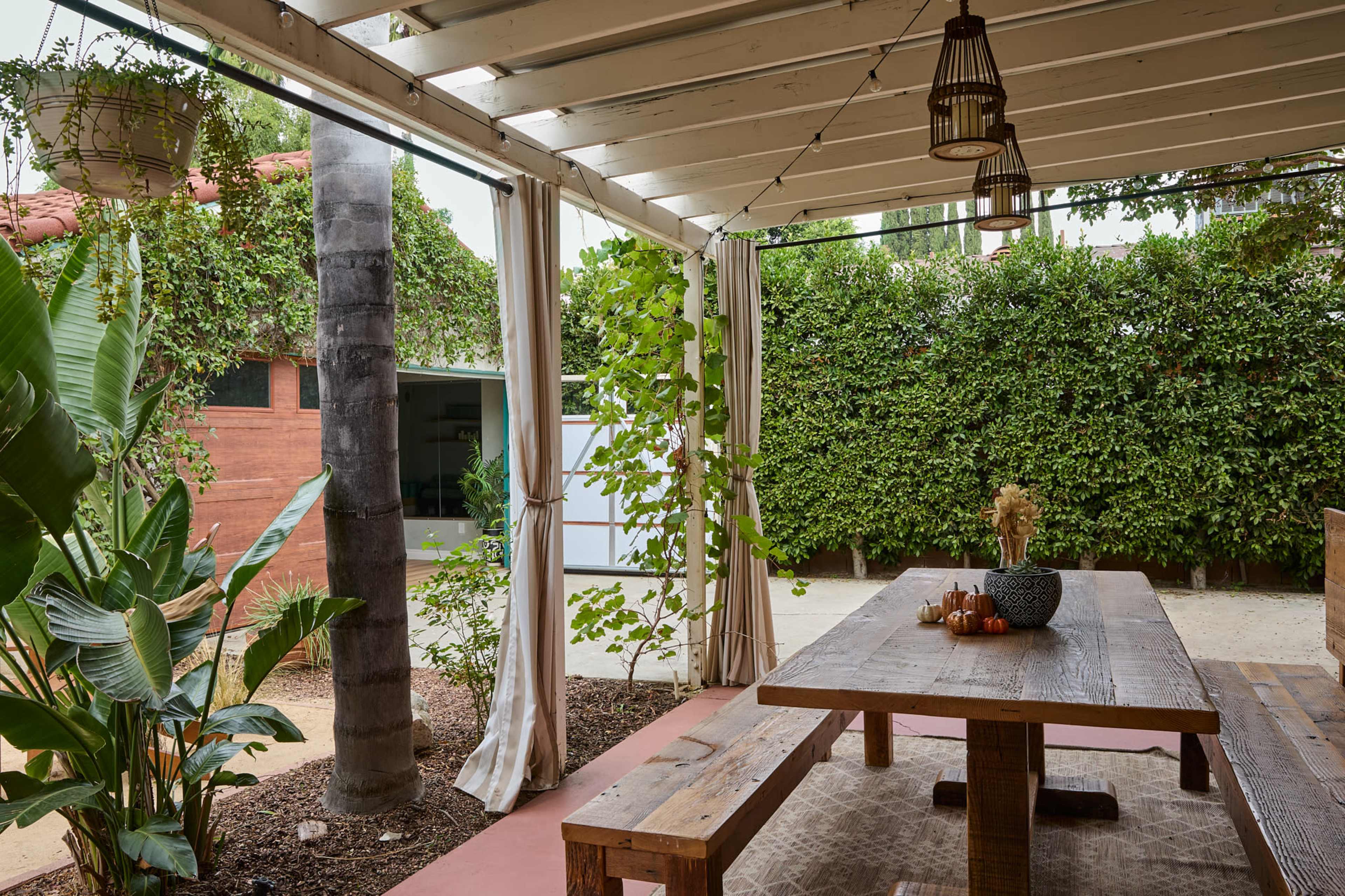The image shows a cozy outdoor dining area with a wooden table, surrounded by greenery and a partially shaded roof.