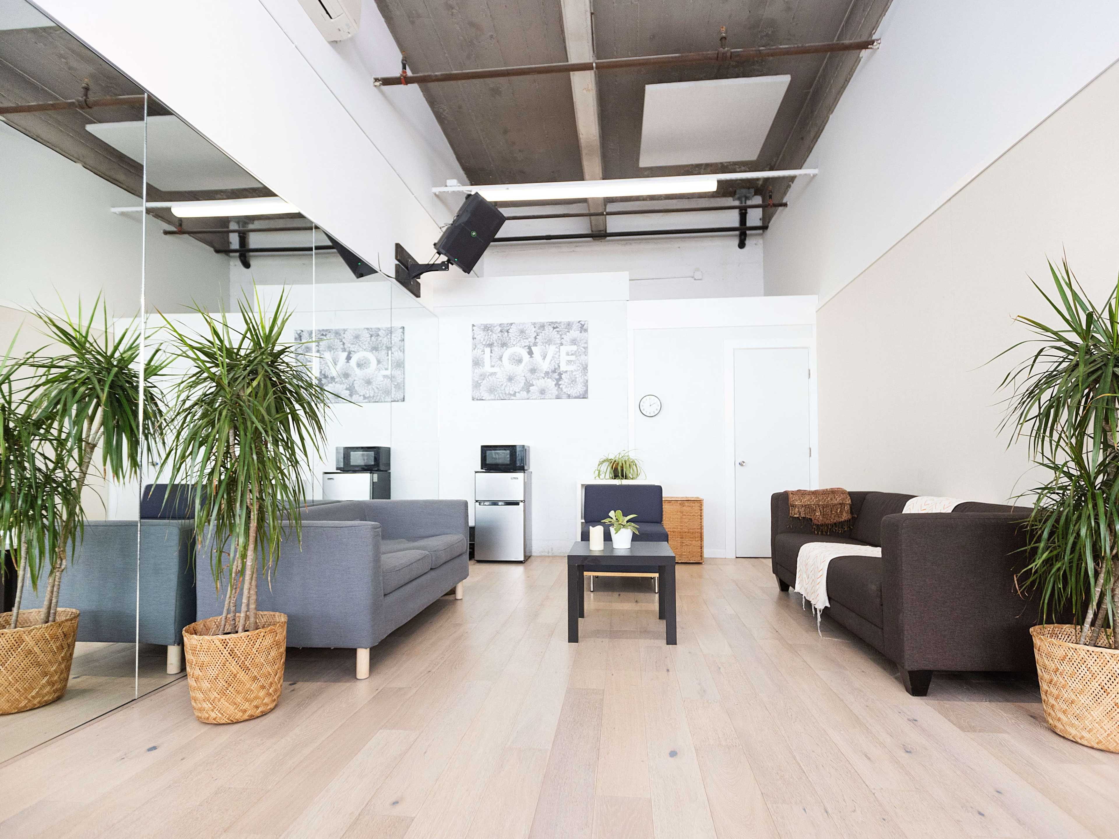 A spacious room features two sofas, a coffee table, potted plants, and wall art, along with a kitchenette area and a clock on the wall.