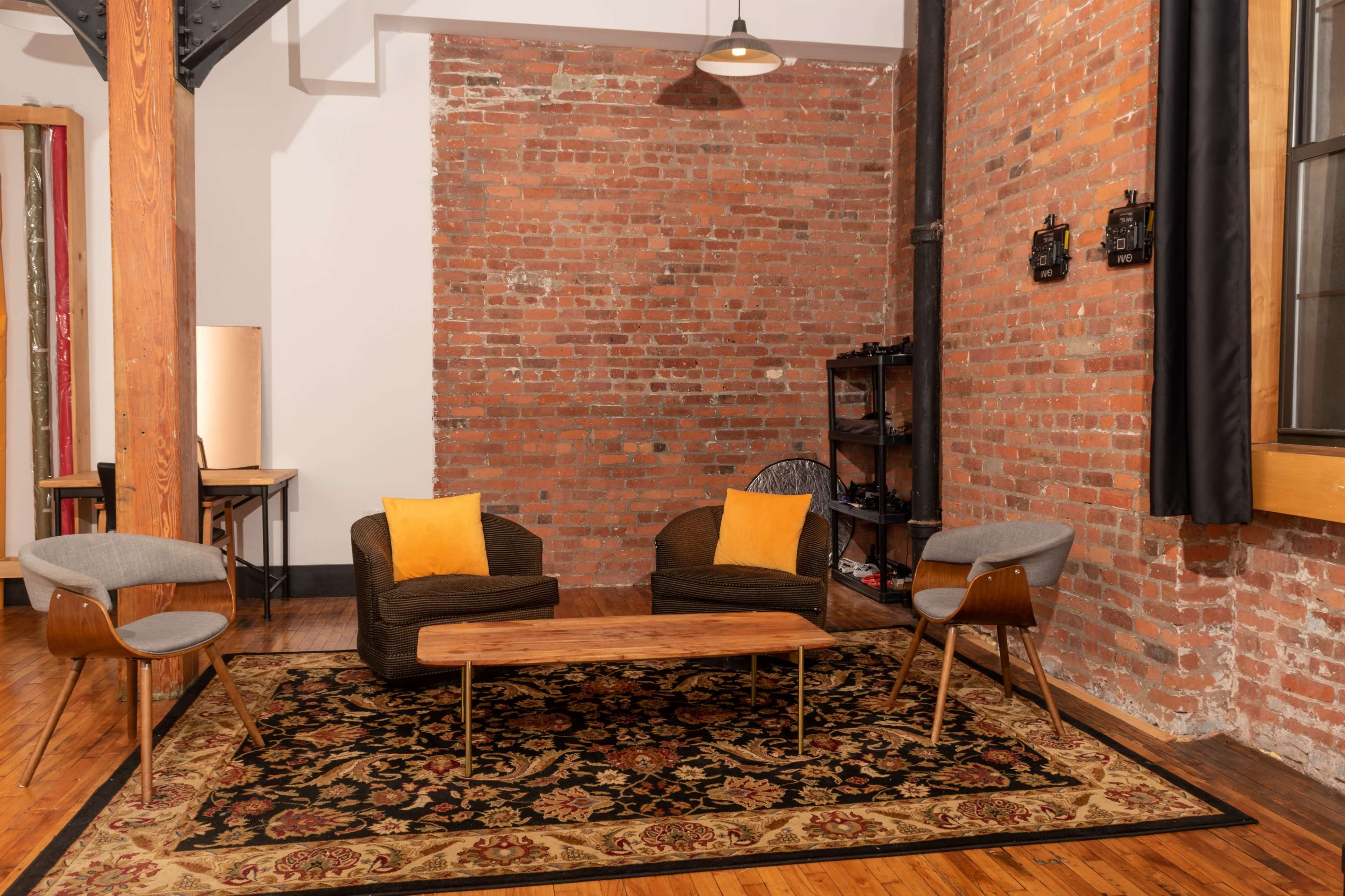 The scene features a living room with two upholstered chairs, a wooden coffee table, and a patterned rug, set against a backdrop of exposed brick walls.