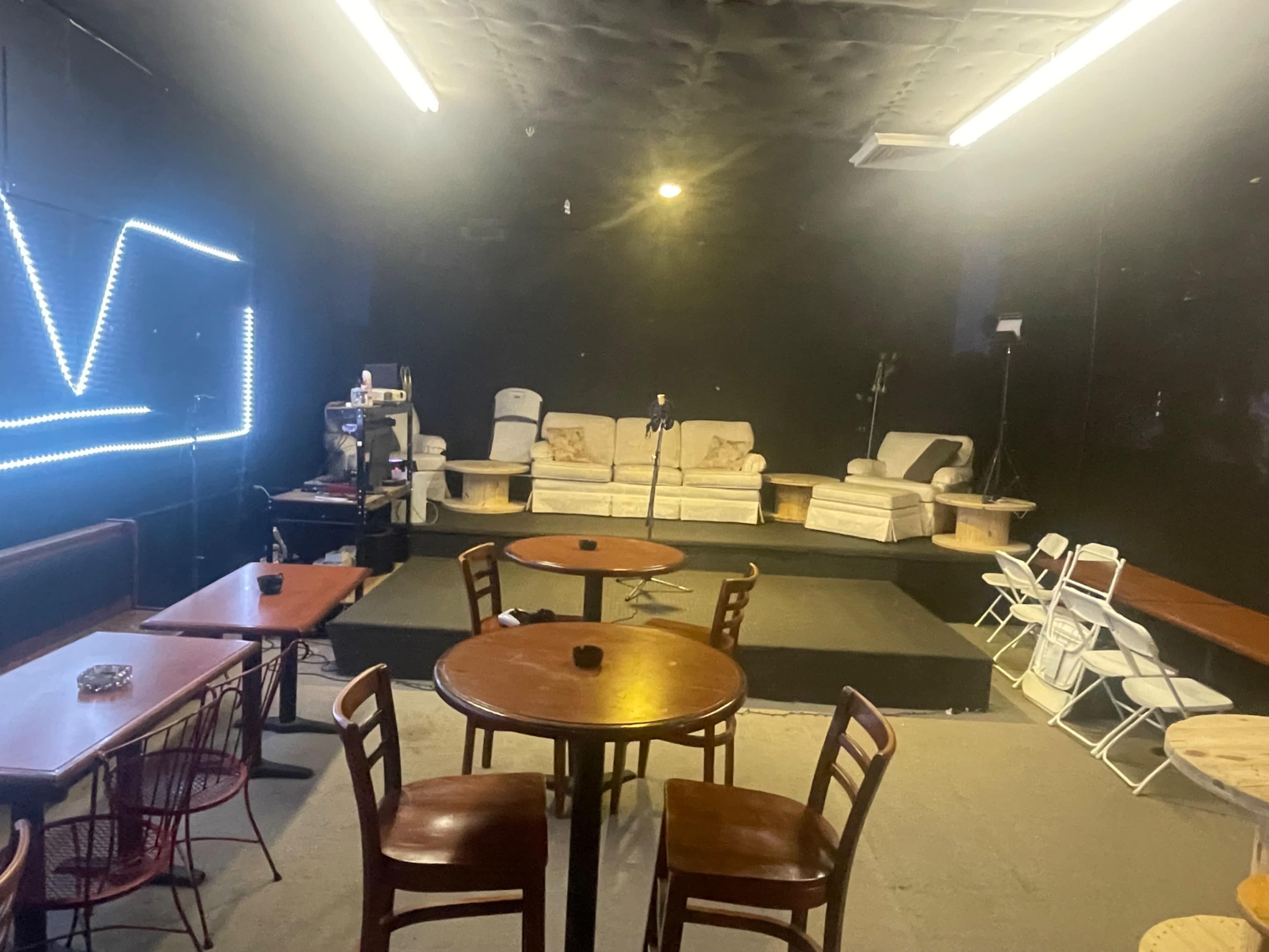 The image shows a dimly lit stage area with a microphone stand, surrounded by tables and chairs in a performance space.