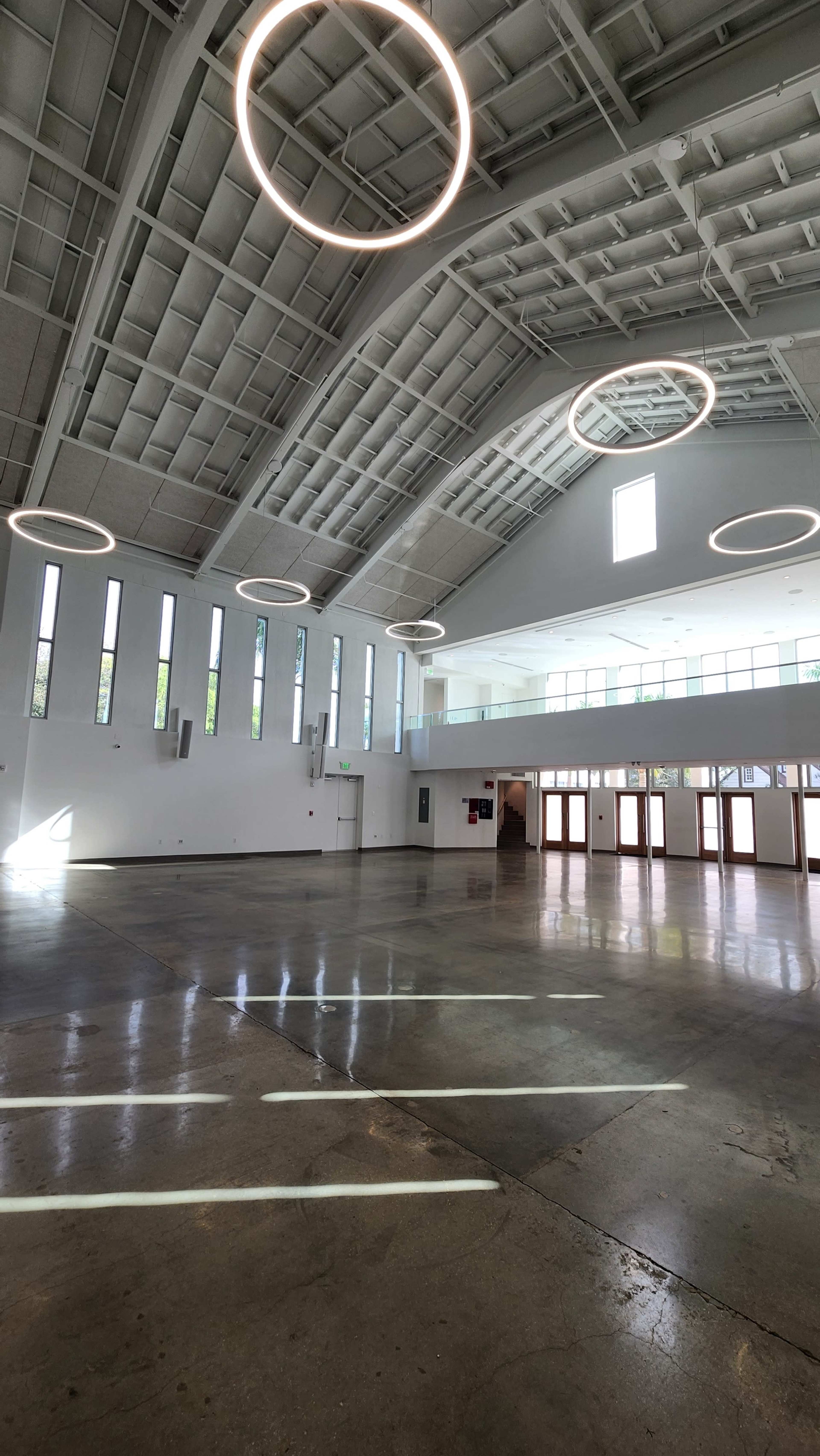 The image shows a spacious, modern interior with a high, arched ceiling and circular light fixtures, featuring large windows and a polished concrete floor.