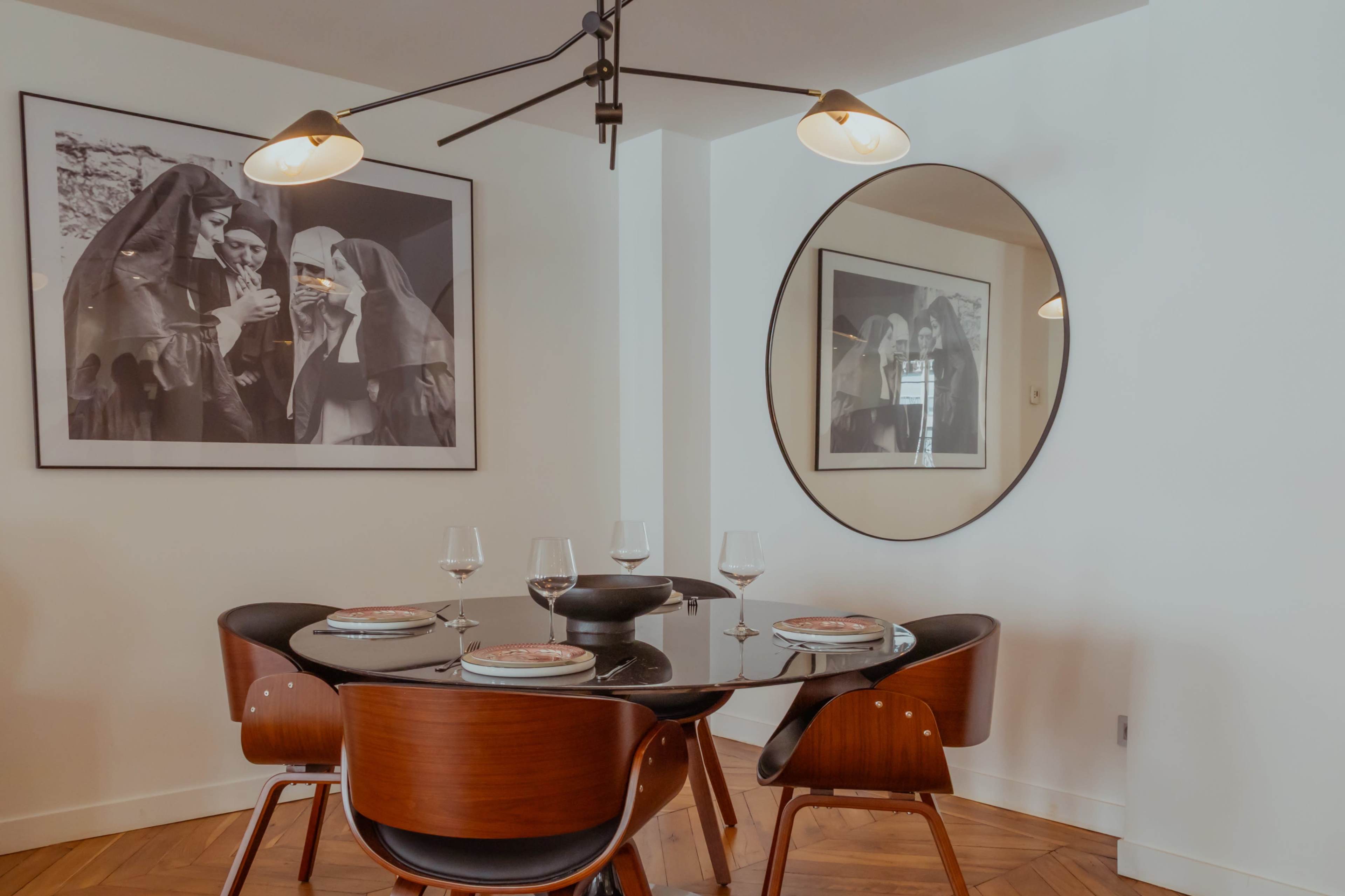 A modern dining area features a round table set with glassware and plates, illuminated by a contemporary light fixture, and a large round mirror reflecting an artwork of two women.