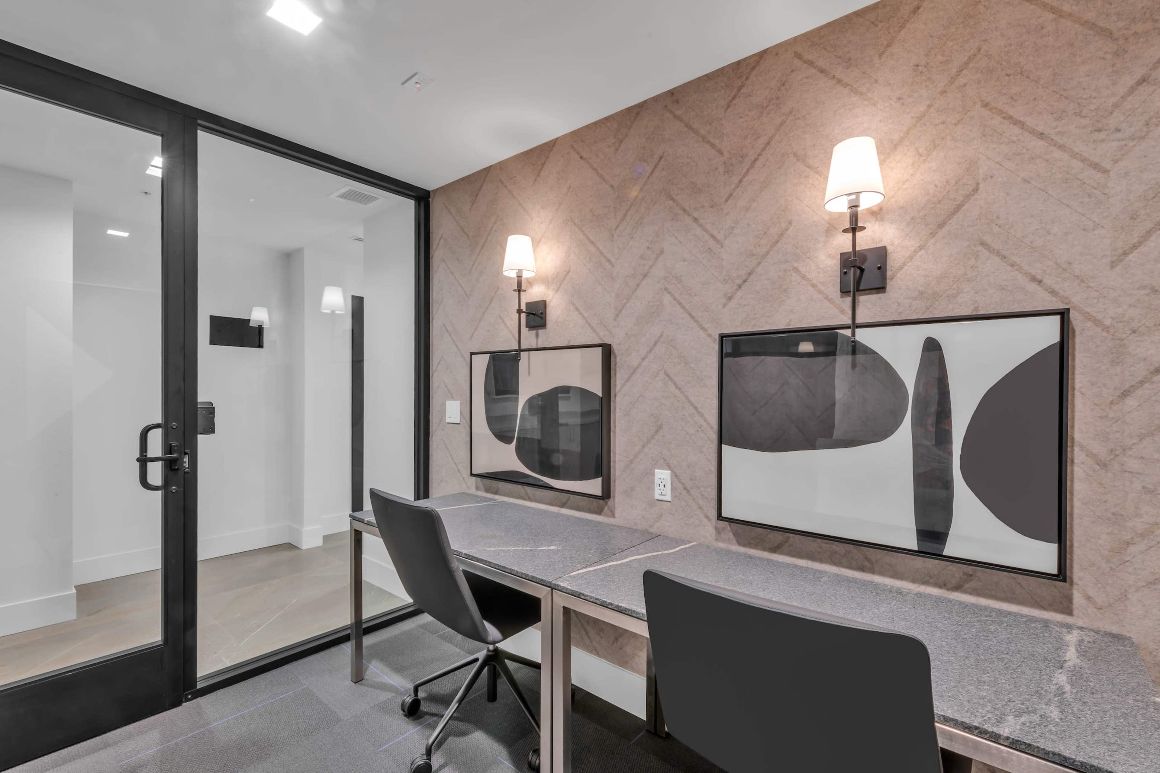 A modern workspace featuring two desks against a textured wall with framed abstract art and wall sconces, alongside a glass door leading to another area.