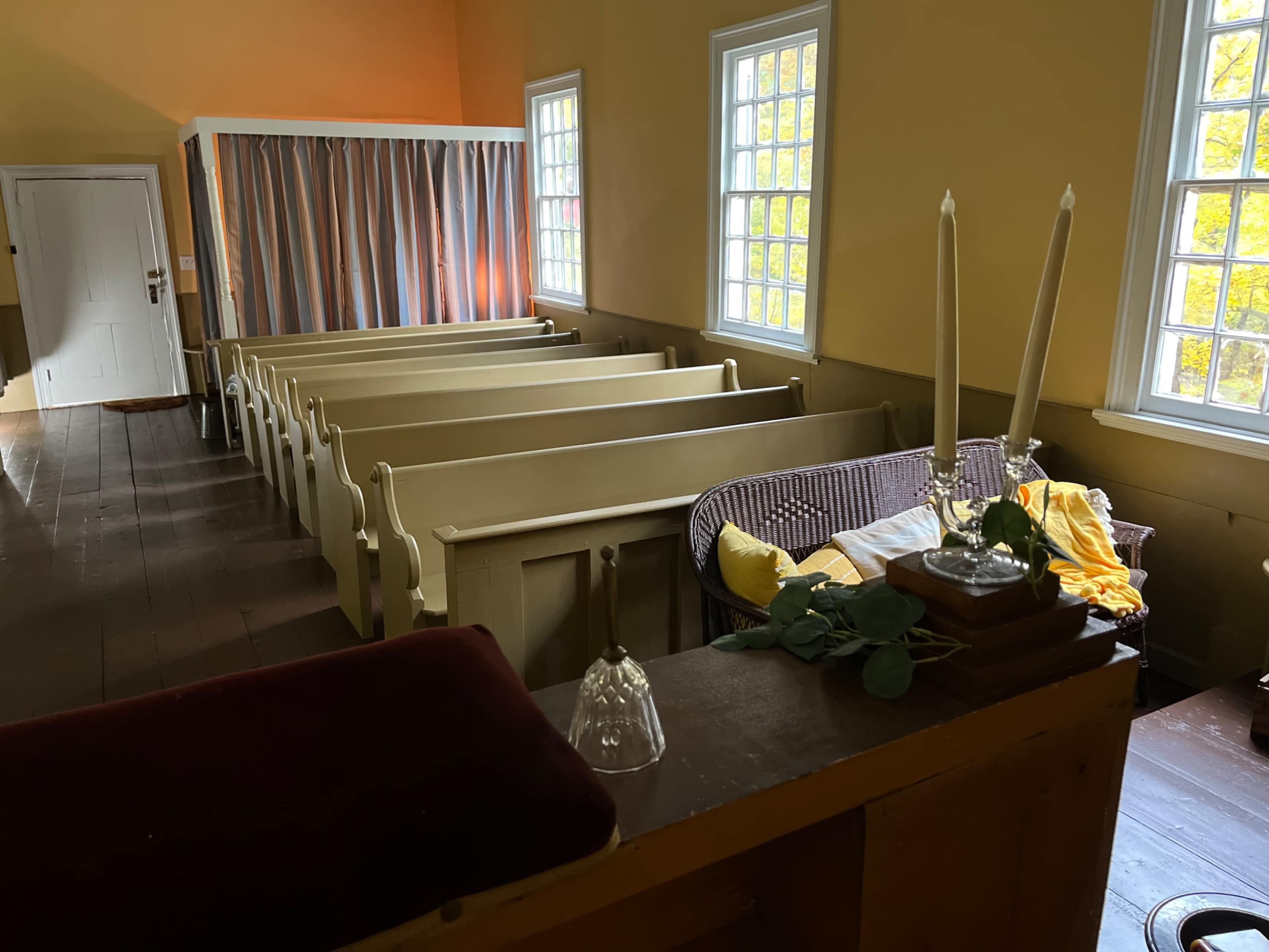 The image depicts the interior of a historic room with several wooden pews arranged neatly, a couch with a yellow throw blanket, and a small table with candles.
