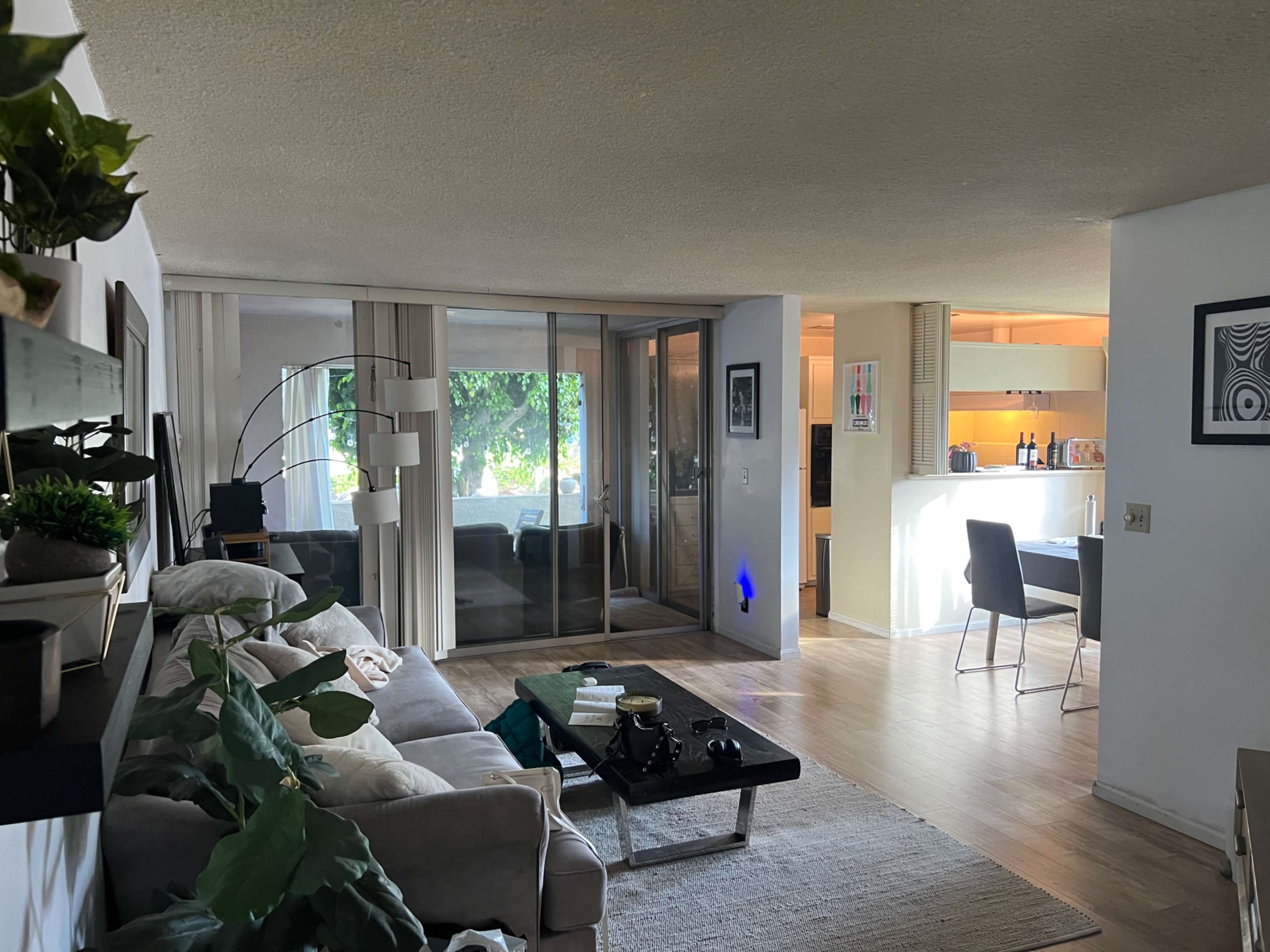 A modern living area with a gray couch, a coffee table, and a kitchen in the background, featuring a sliding glass door that opens to a patio.