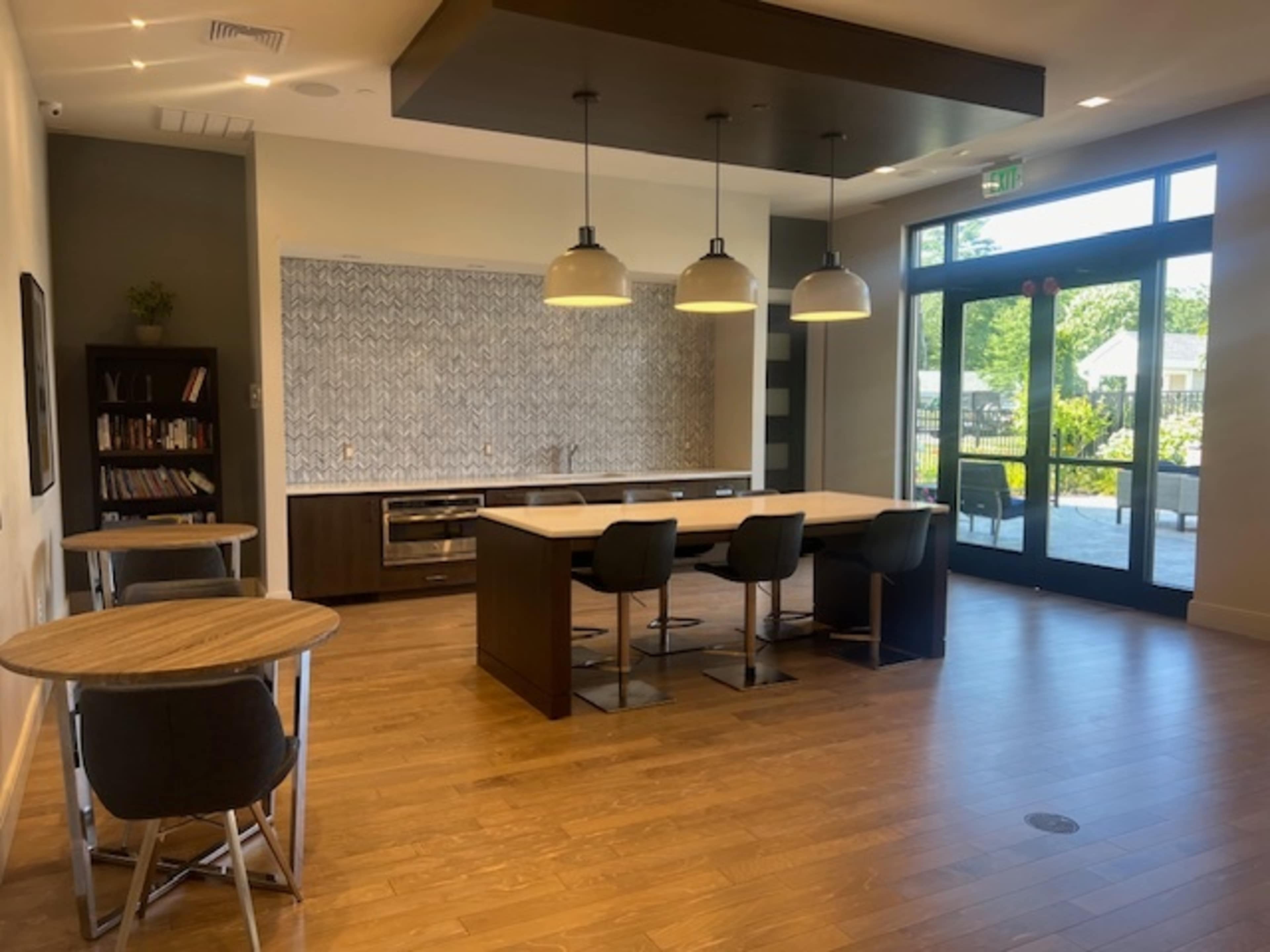 A modern kitchen space features an island with bar stools, a wall of textured tiles, and a dining table positioned nearby, with large windows allowing natural light.